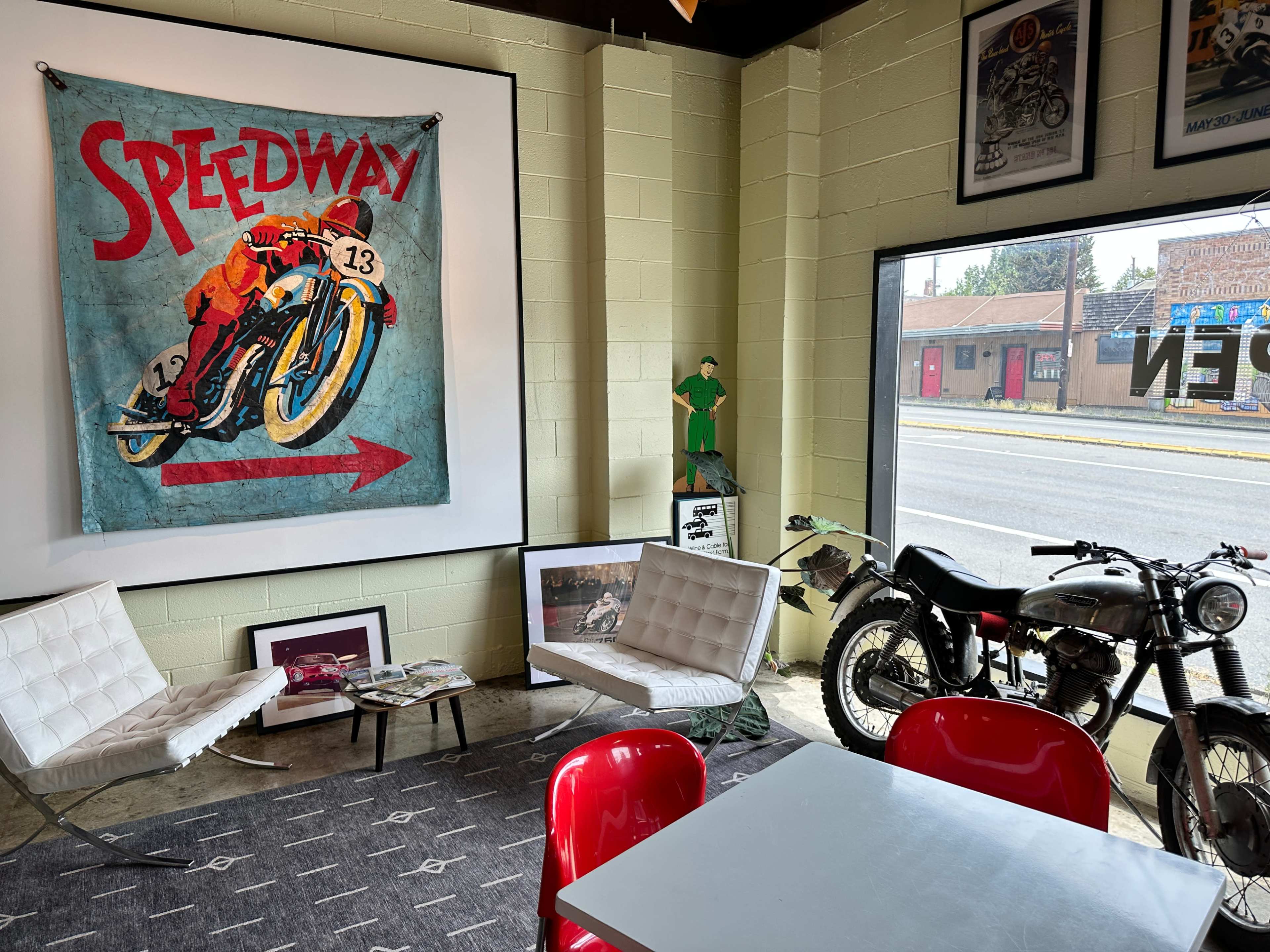 The image shows a vintage-themed room featuring a large "Speedway" mural, two modern white chairs, a small gray table, various framed pictures on the walls, and a classic motorcycle parked beside a window.