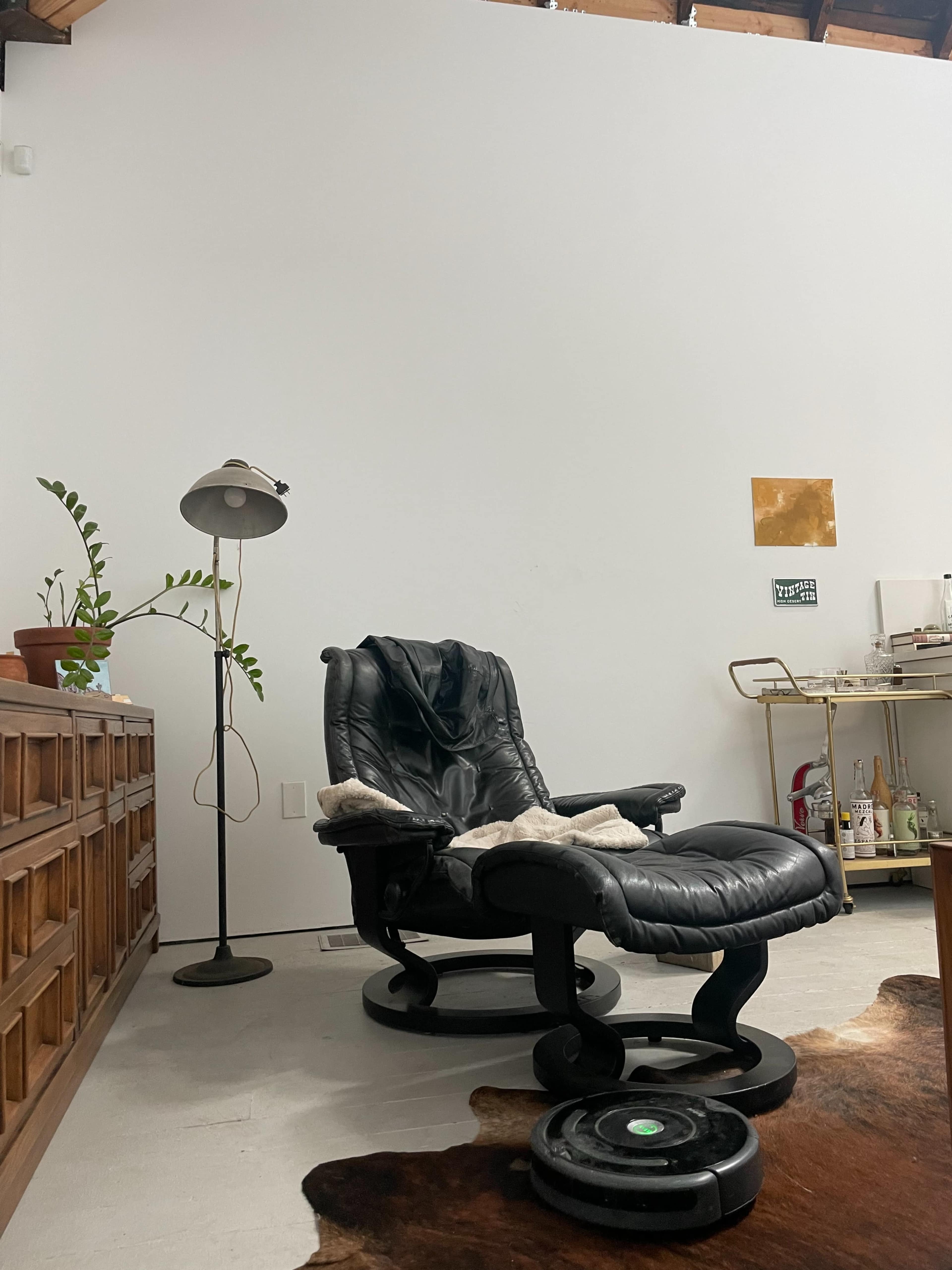 A black recliner chair sits next to a floor lamp and a wooden cabinet in a minimalist room.