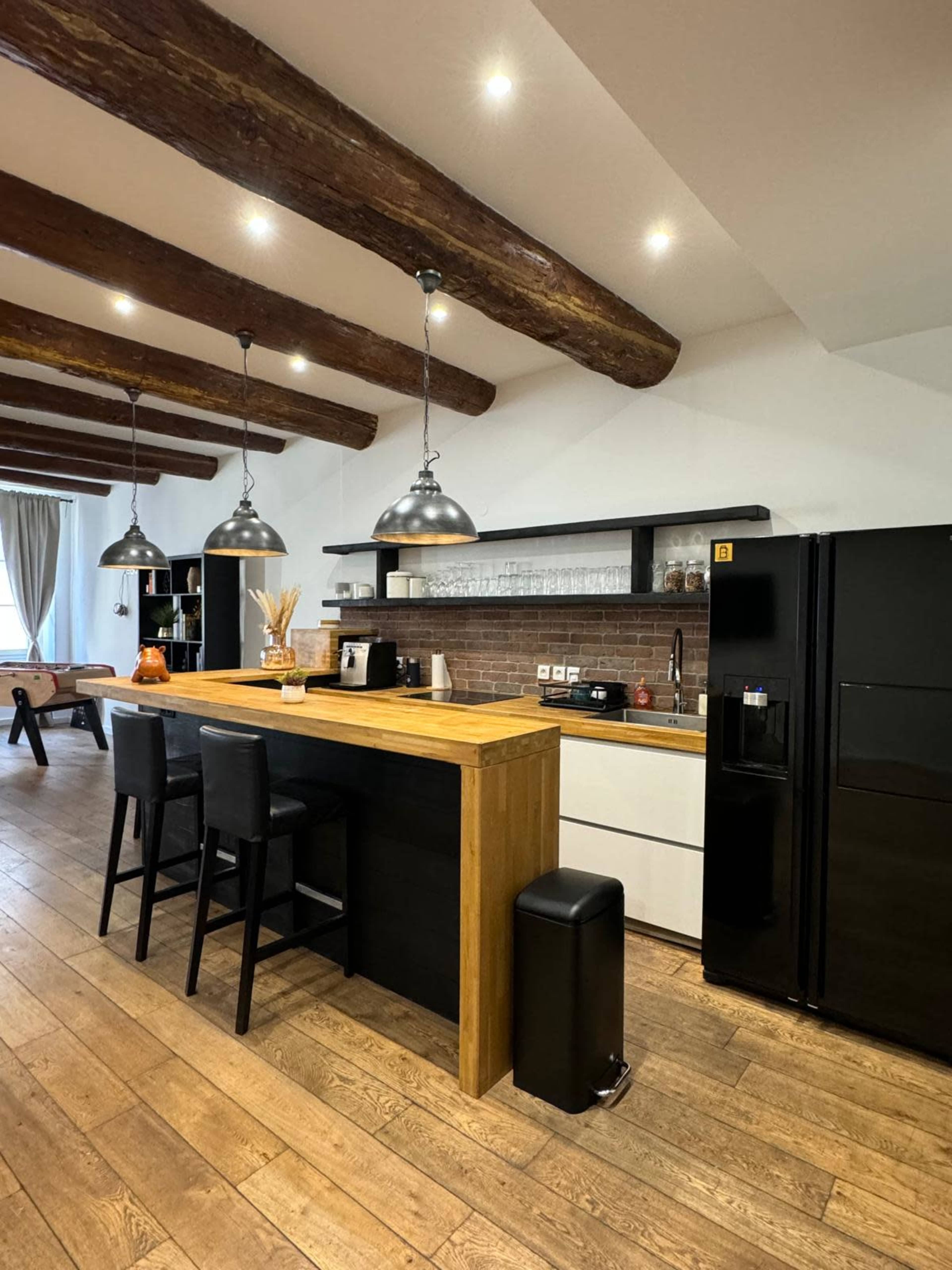 The kitchen features a wooden countertop with three black bar stools, industrial-style pendant lights, and a mixture of modern appliances against a brick wall.