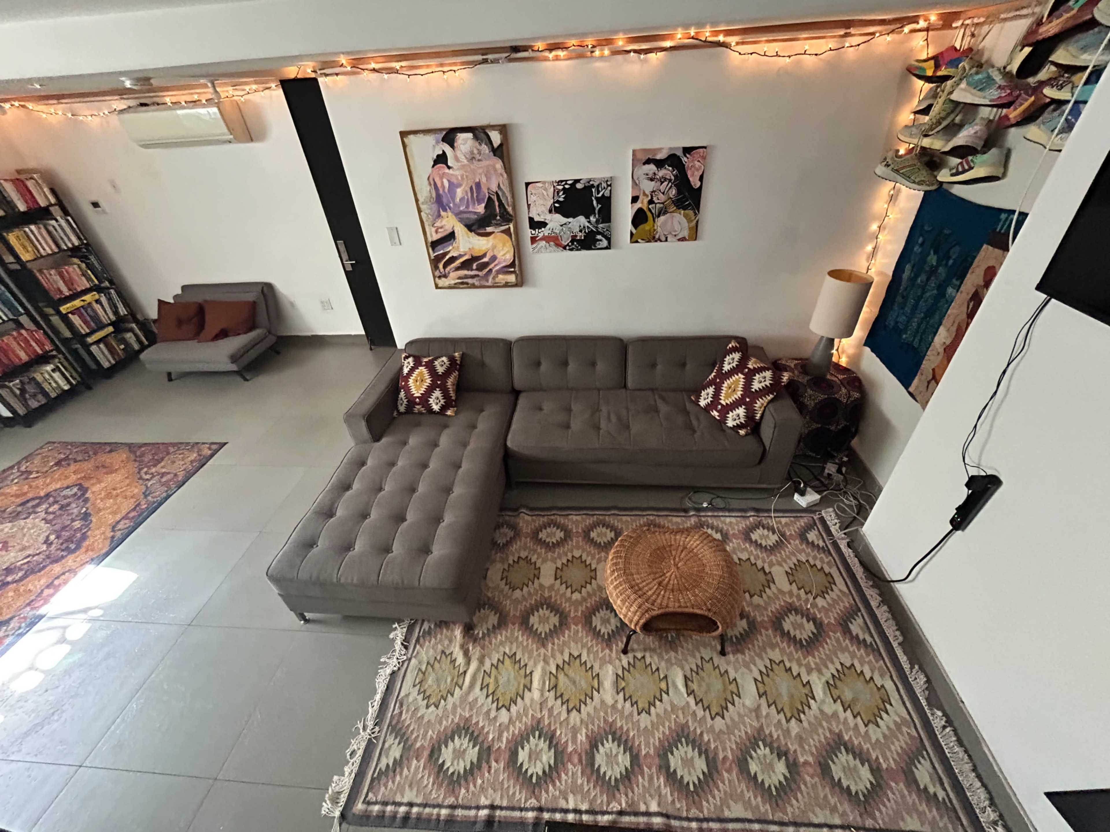 The image shows a modern living room with a gray sectional sofa, a patterned rug, a woven coffee table, and various artworks displayed on the walls.