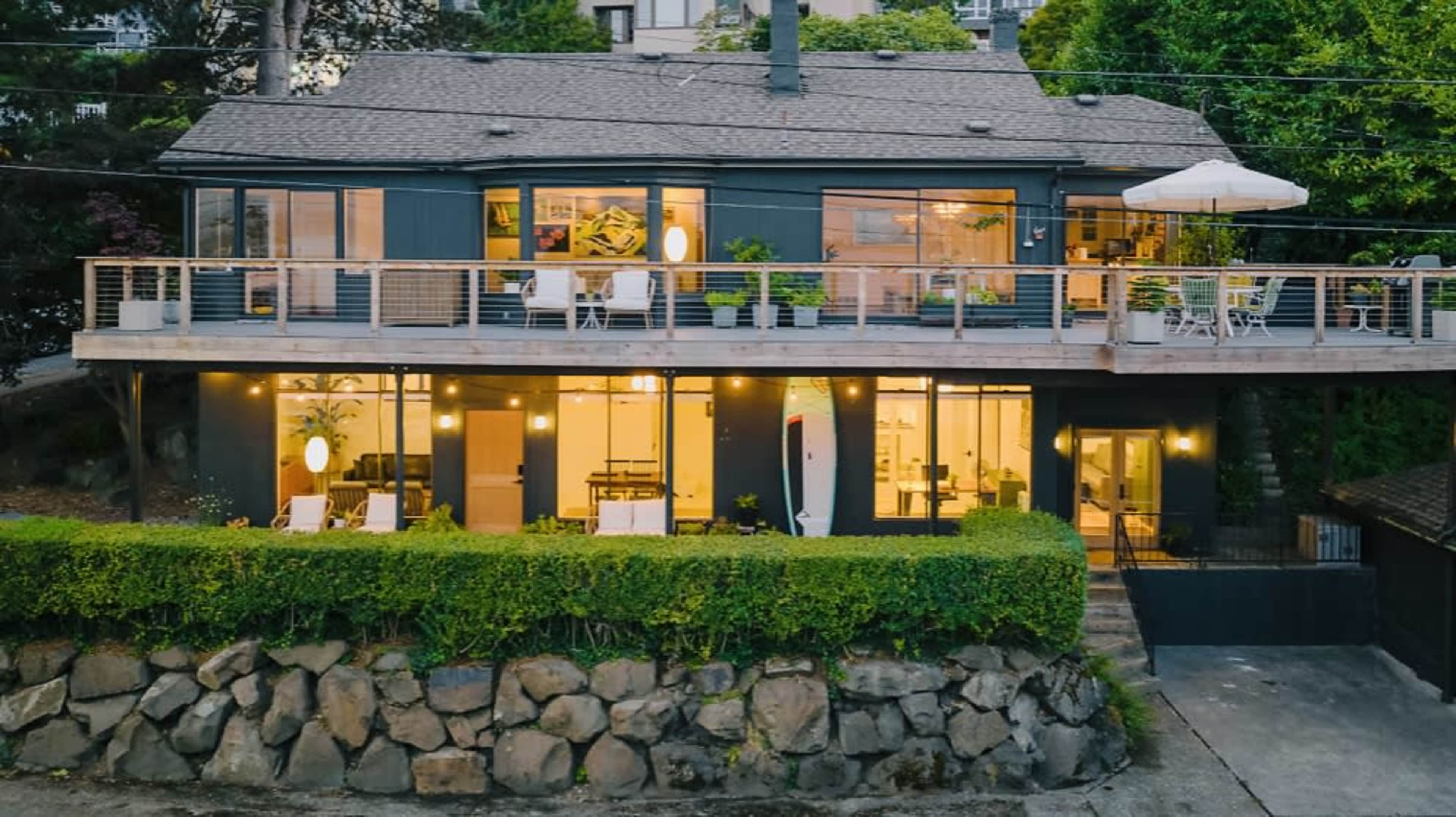 A modern two-story house with large windows and a deck is surrounded by greenery and features a stone wall and outdoor seating.
