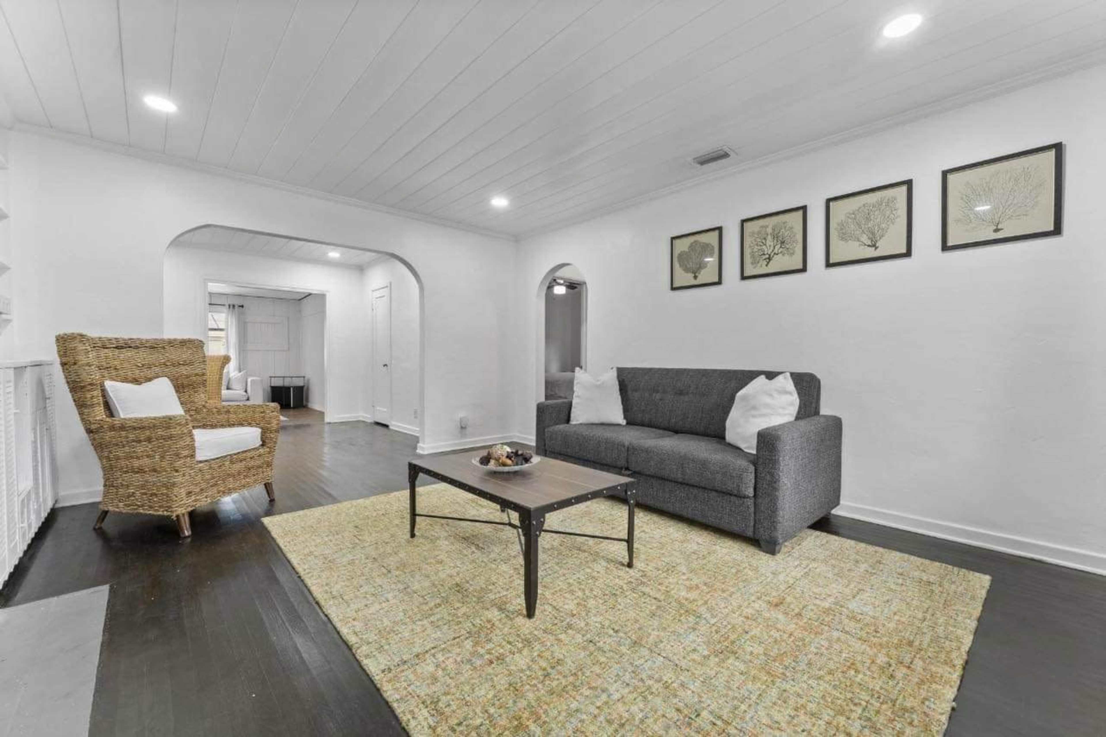 A bright living room with a gray sofa, a coffee table, and a woven chair, featuring framed artwork on the wall.