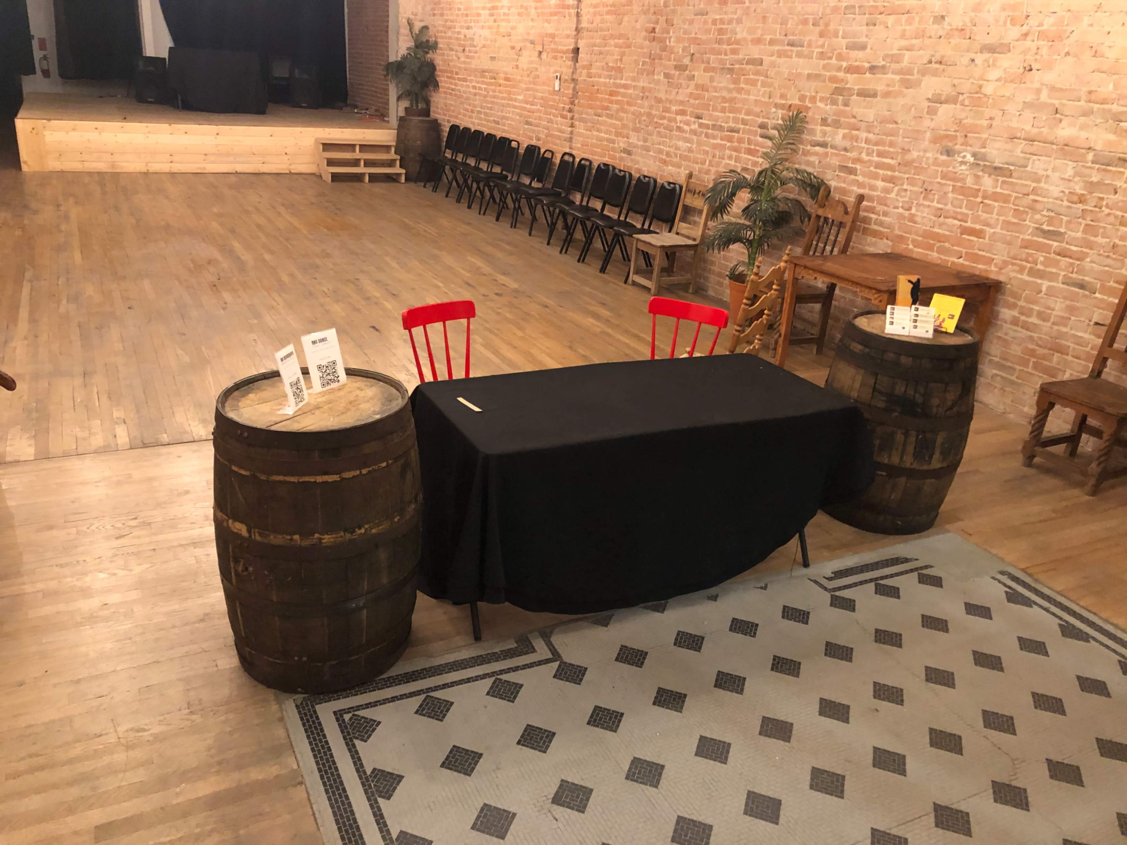 A rustic event space with two barrel tables, a black tablecloth, and red chairs, set against a brick wall and wooden floor.