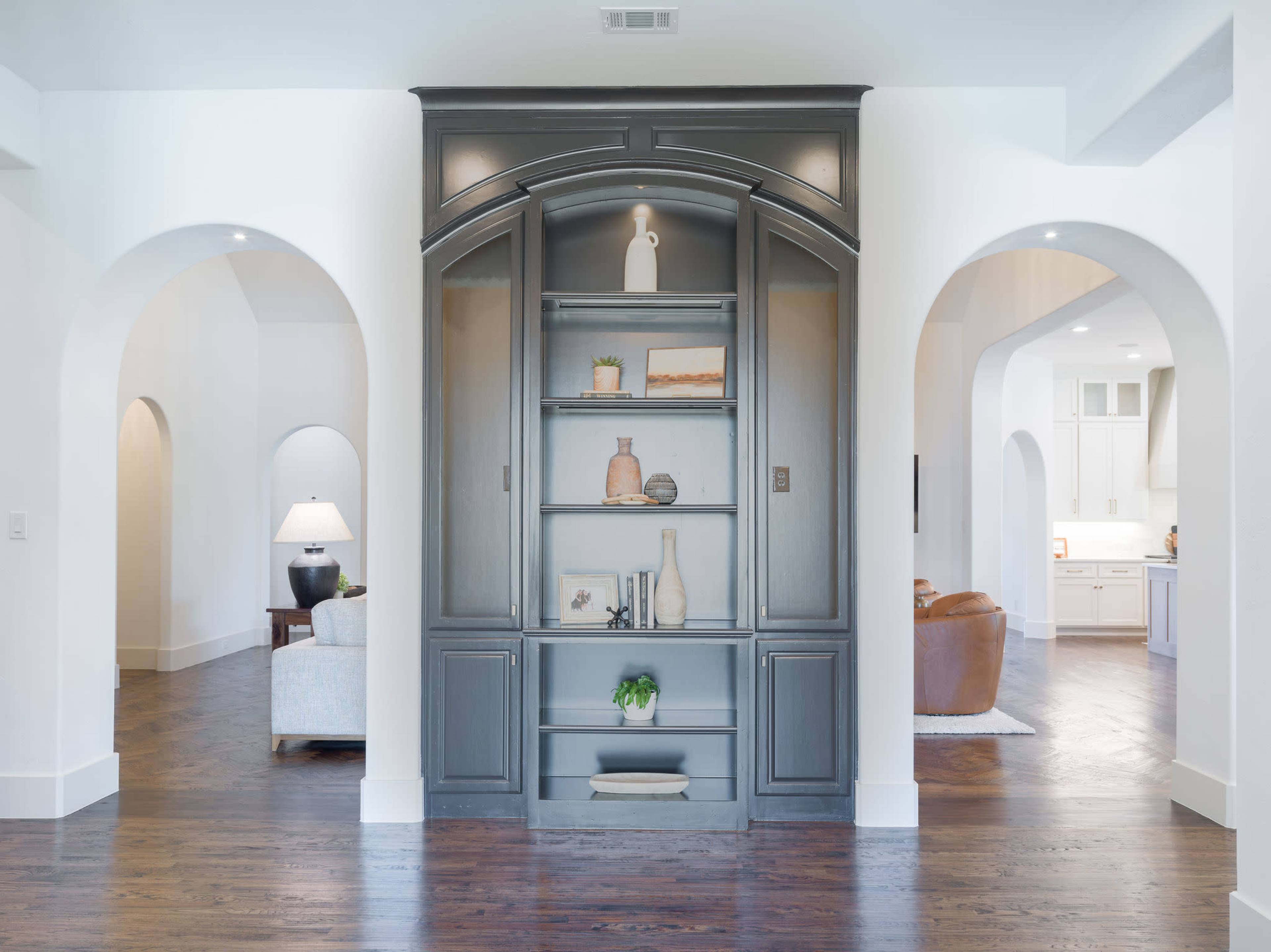 A gray built-in shelving unit stands between two archways in a bright, open living space with light wood flooring.