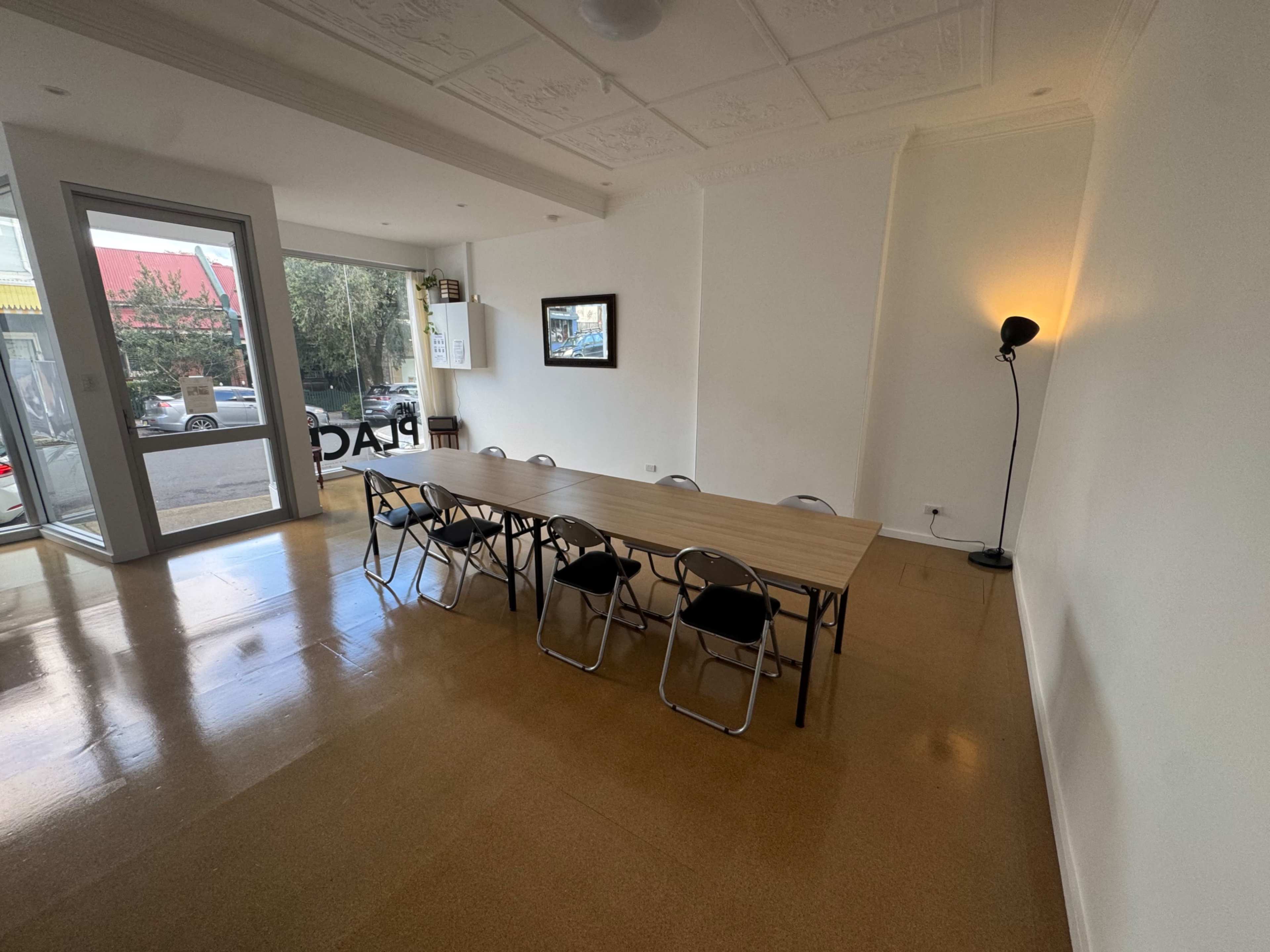 A long wooden table with chairs sits in a bright, minimalist room featuring large windows and a standing lamp.