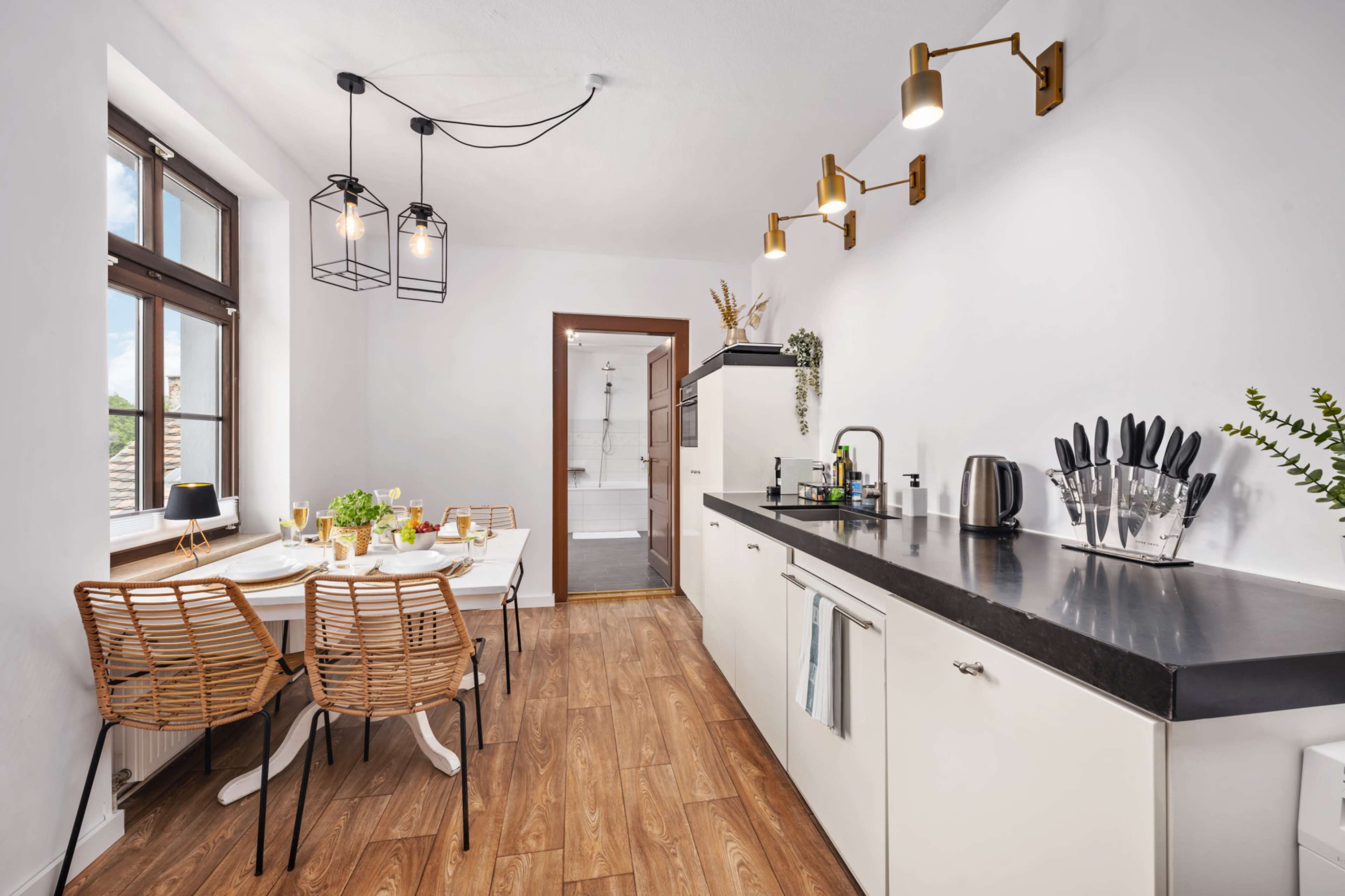 The image shows a modern kitchen with a white and black color scheme, featuring wooden flooring, a dining table with chairs, and a doorway leading to a bathroom.