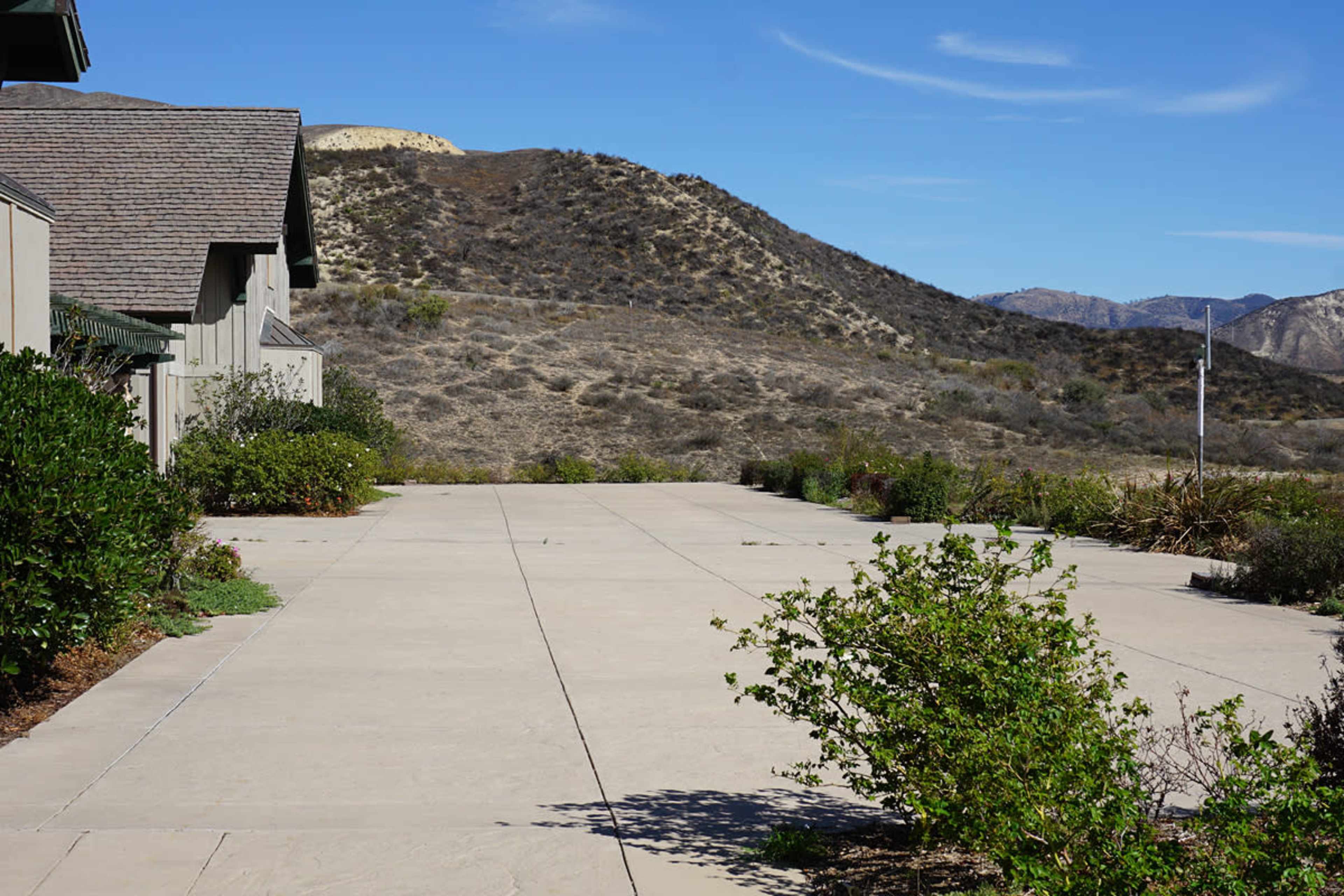A concrete pathway leads through landscaped bushes towards a hilly area under a clear blue sky.