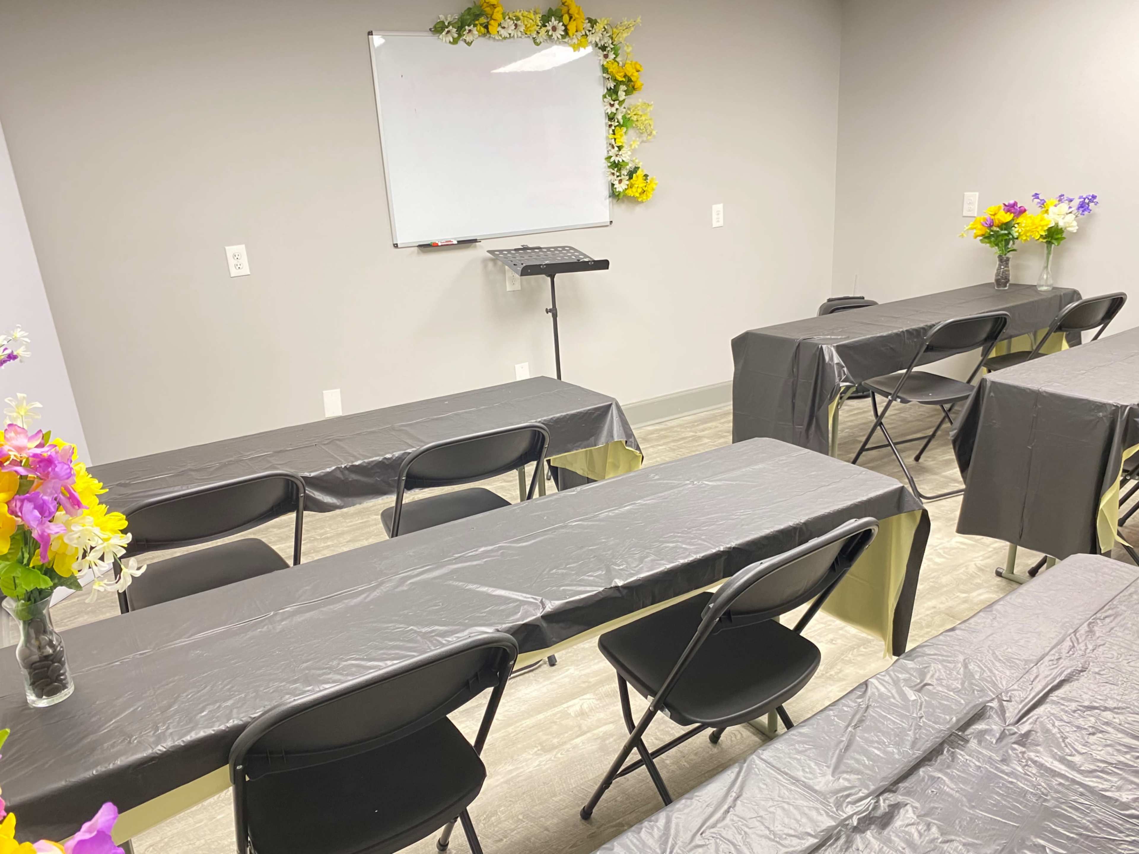 A classroom setup with black tables and chairs, a podium, and floral decorations on each table, featuring a whiteboard at the front.