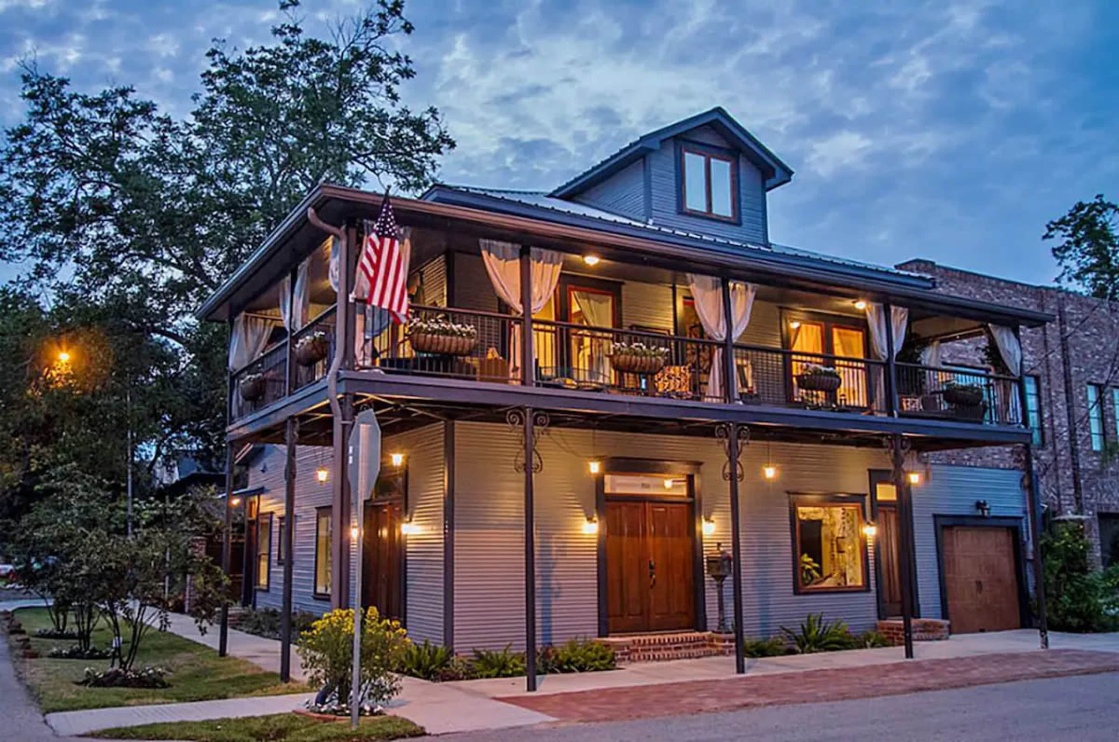 A two-story house with a blue exterior, balconies, and an American flag displayed, surrounded by trees and outdoor lighting.