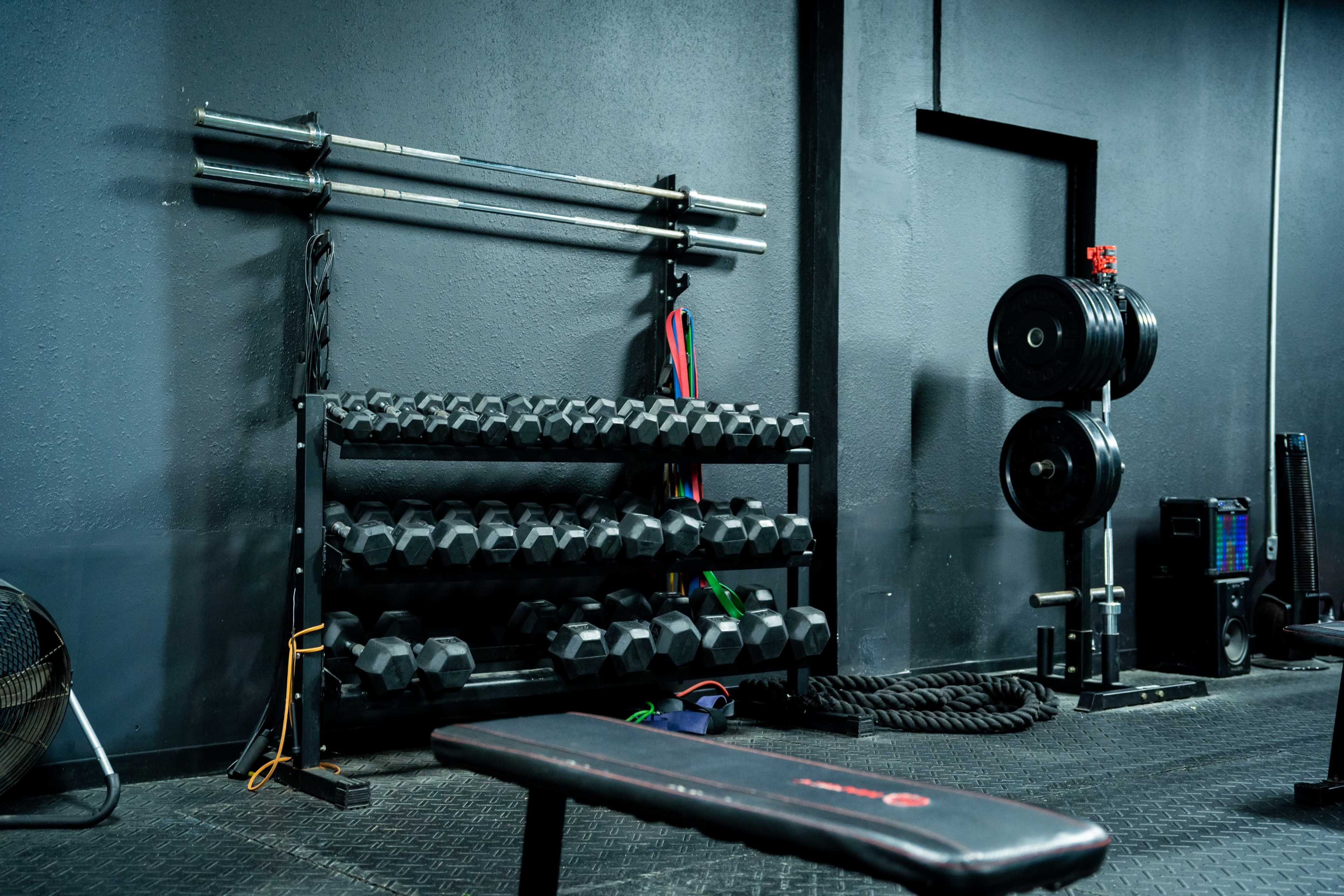 A gym interior featuring a rack of dumbbells, a barbell, weight plates, a weightlifting bench, and exercise equipment against a dark wall.