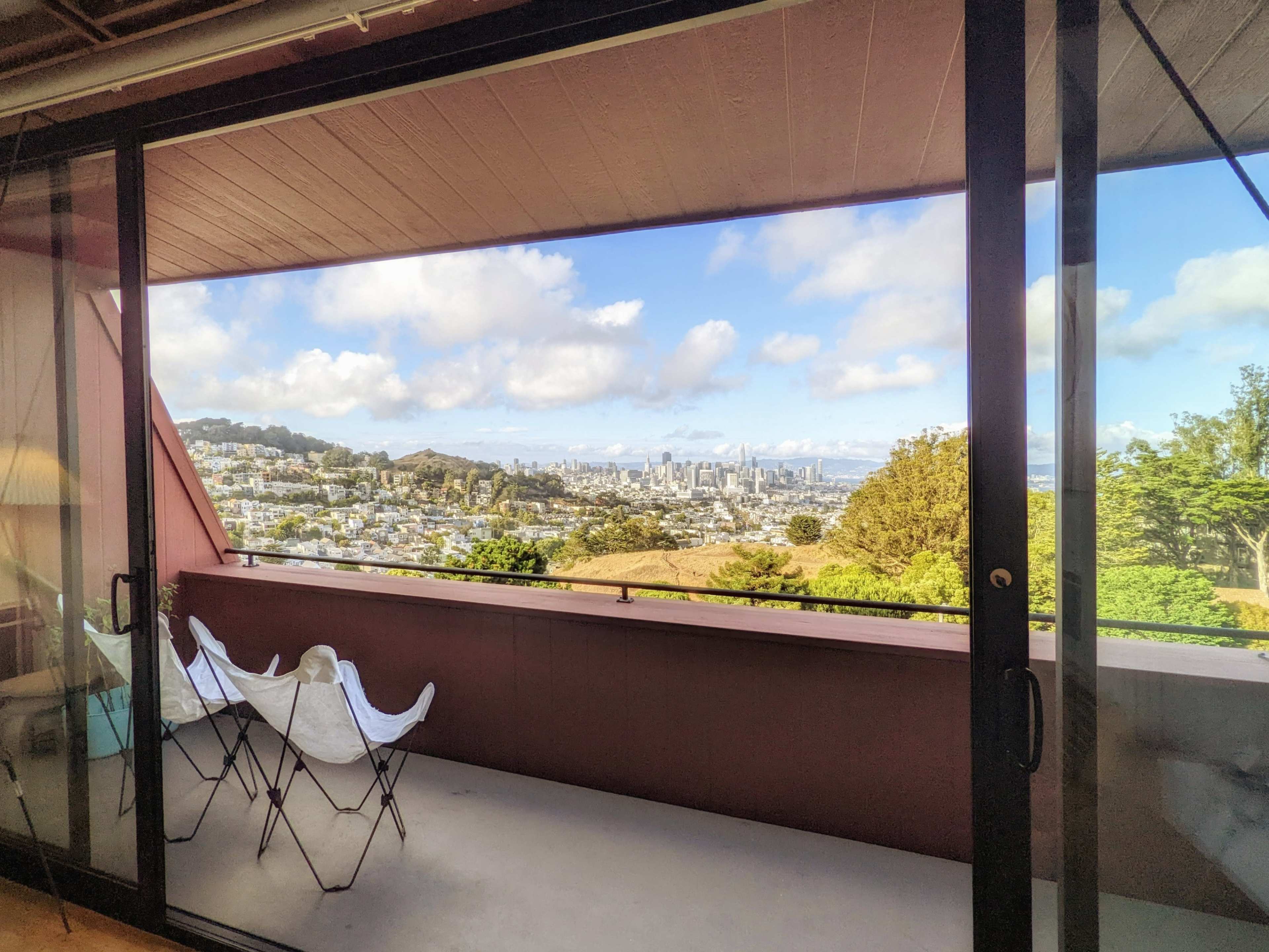 The image shows a view from a balcony with two folding chairs overlooking a city skyline and hills under a partly cloudy sky.