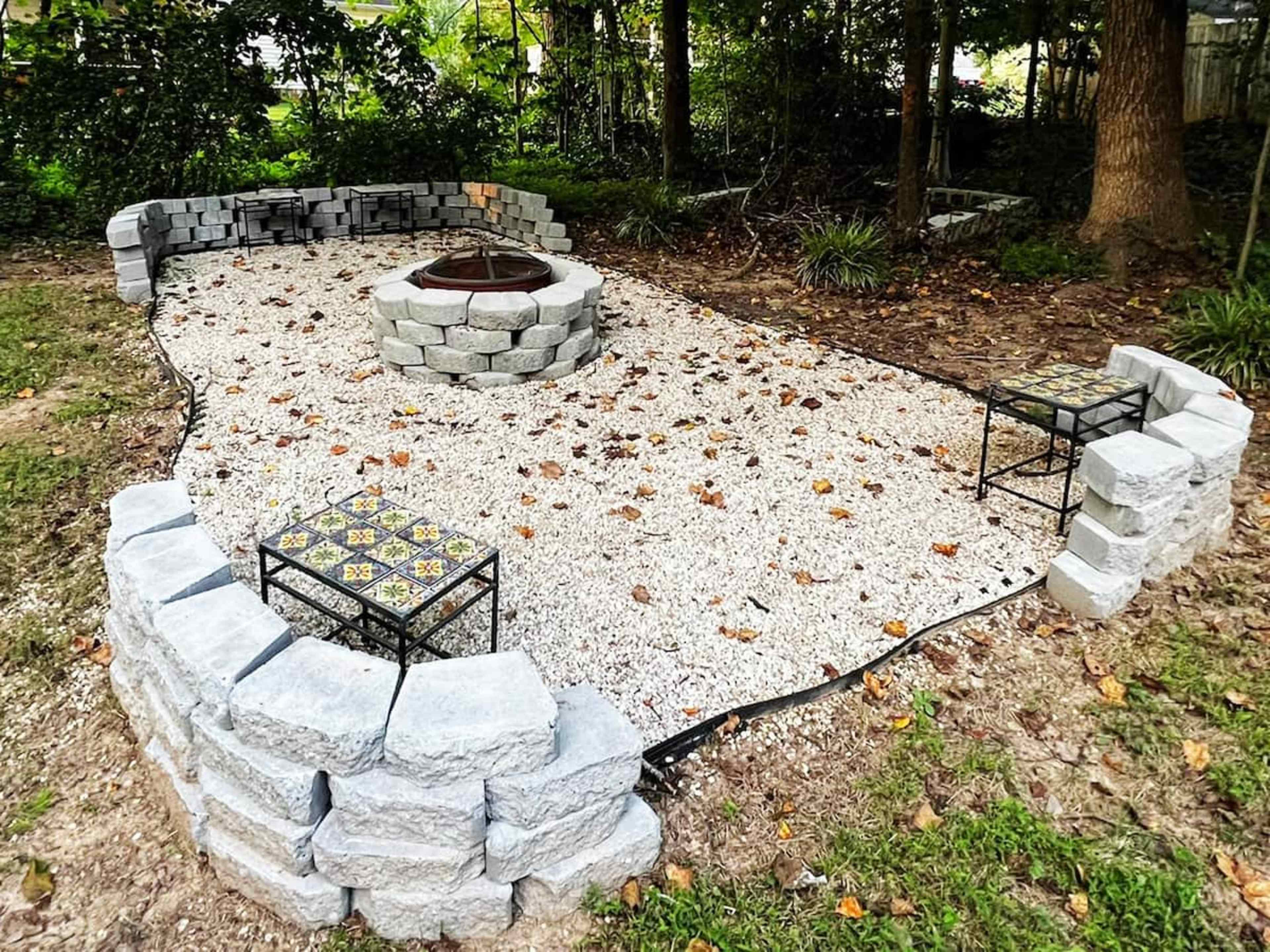 A gravel fire pit area surrounded by a circular stone wall, with two small tables nearby and scattered leaves on the ground.