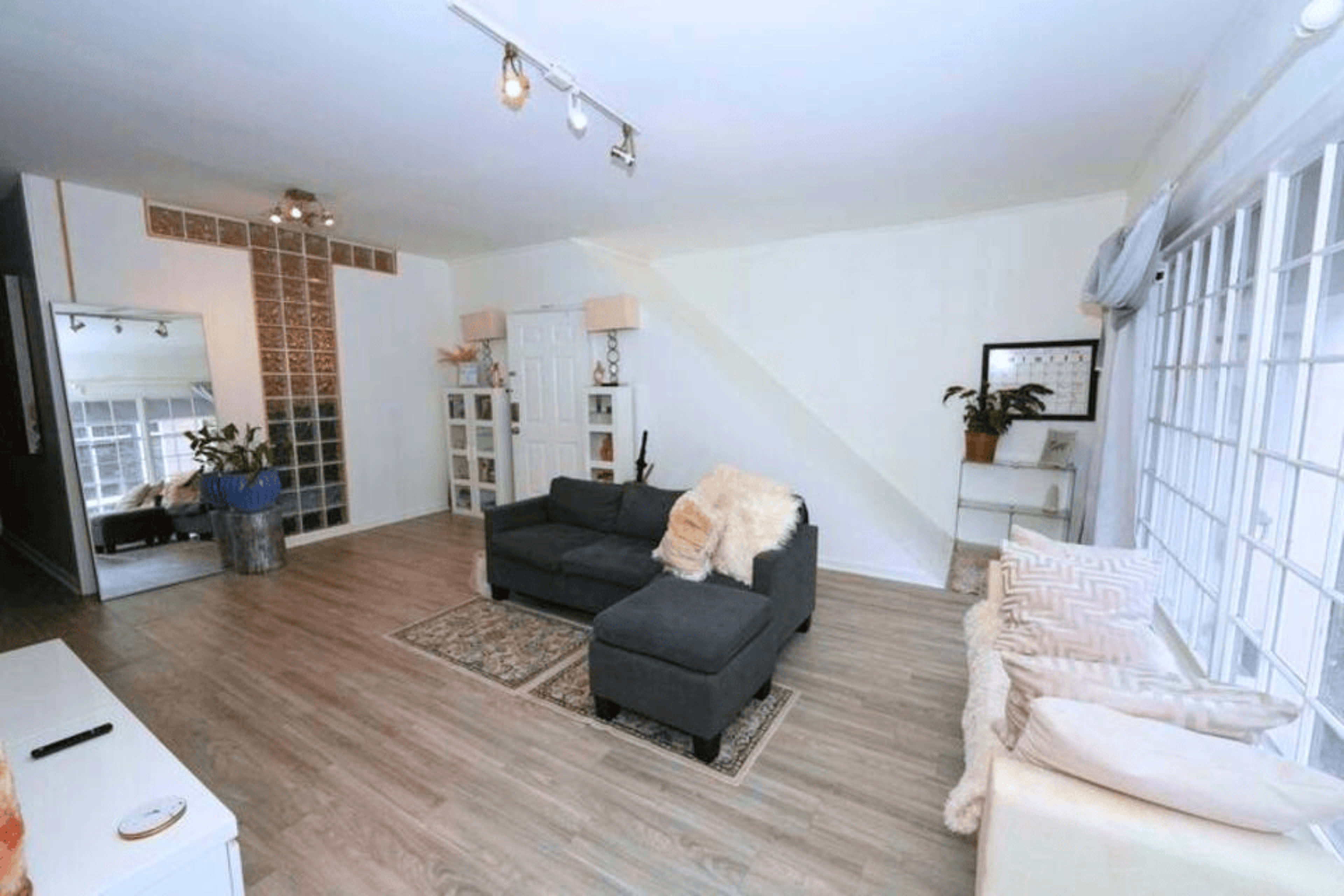 The image shows a spacious living room with a gray sectional sofa, a patterned rug, and large windows allowing natural light.