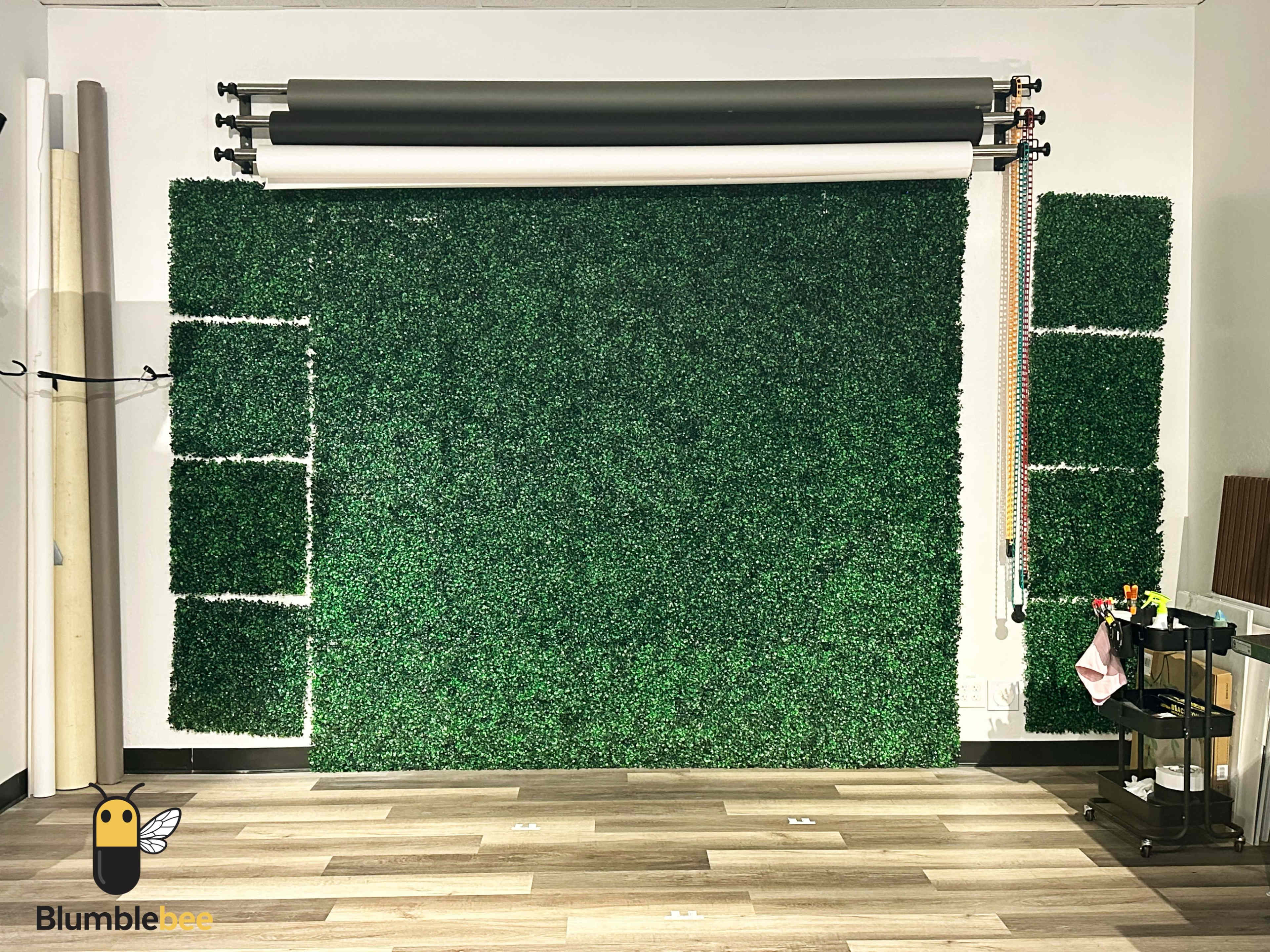 The image shows a wall covered with artificial green grass panels, flanked by a backdrop support system and various tools on a rolling cart.