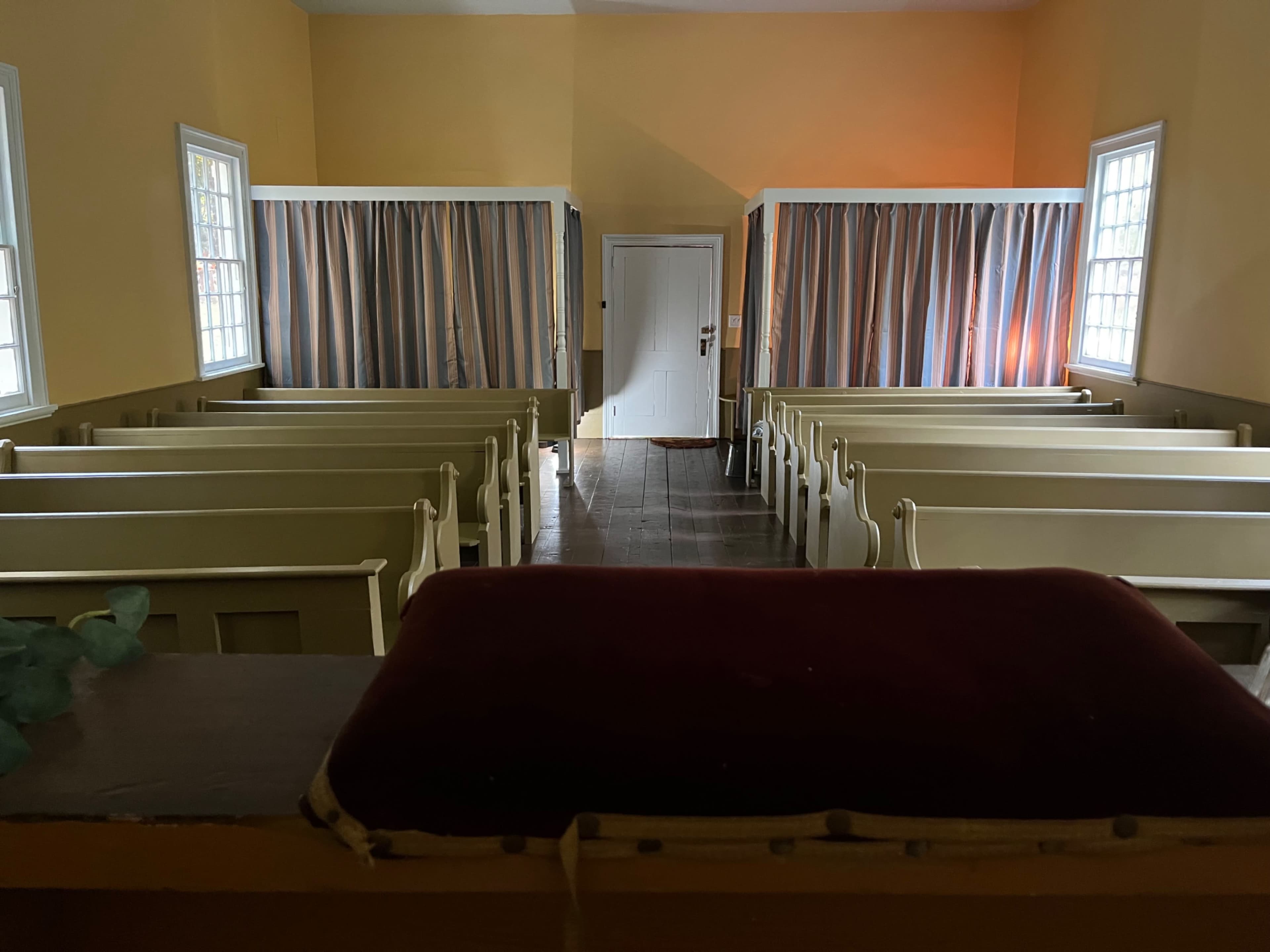 The image shows the interior of a small, empty hall with light yellow walls, featuring rows of pews and curtained sections at each end.