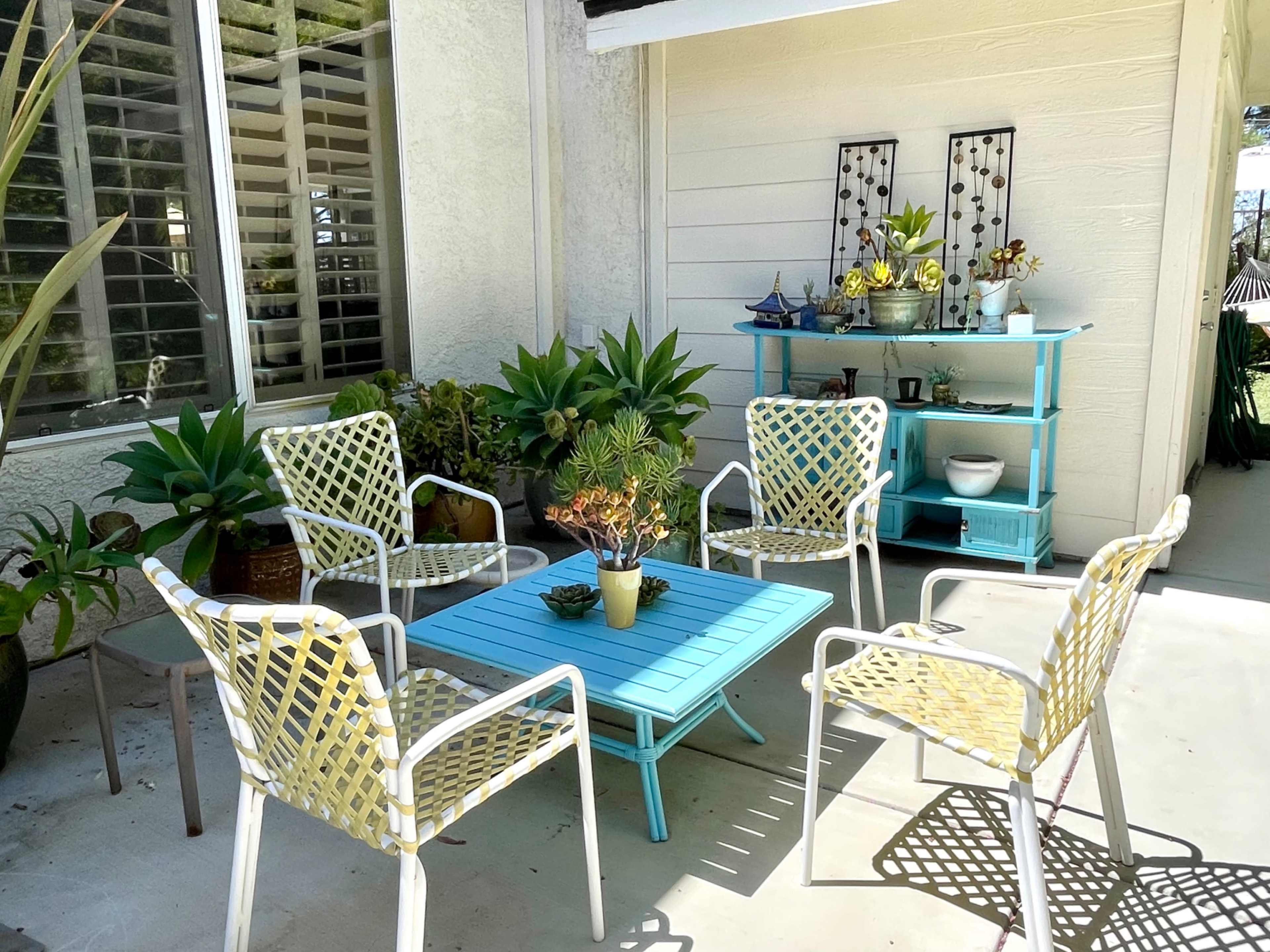 A patio setting features a light blue table surrounded by four chairs with yellow and white patterns, with plants and decorative items in the background.