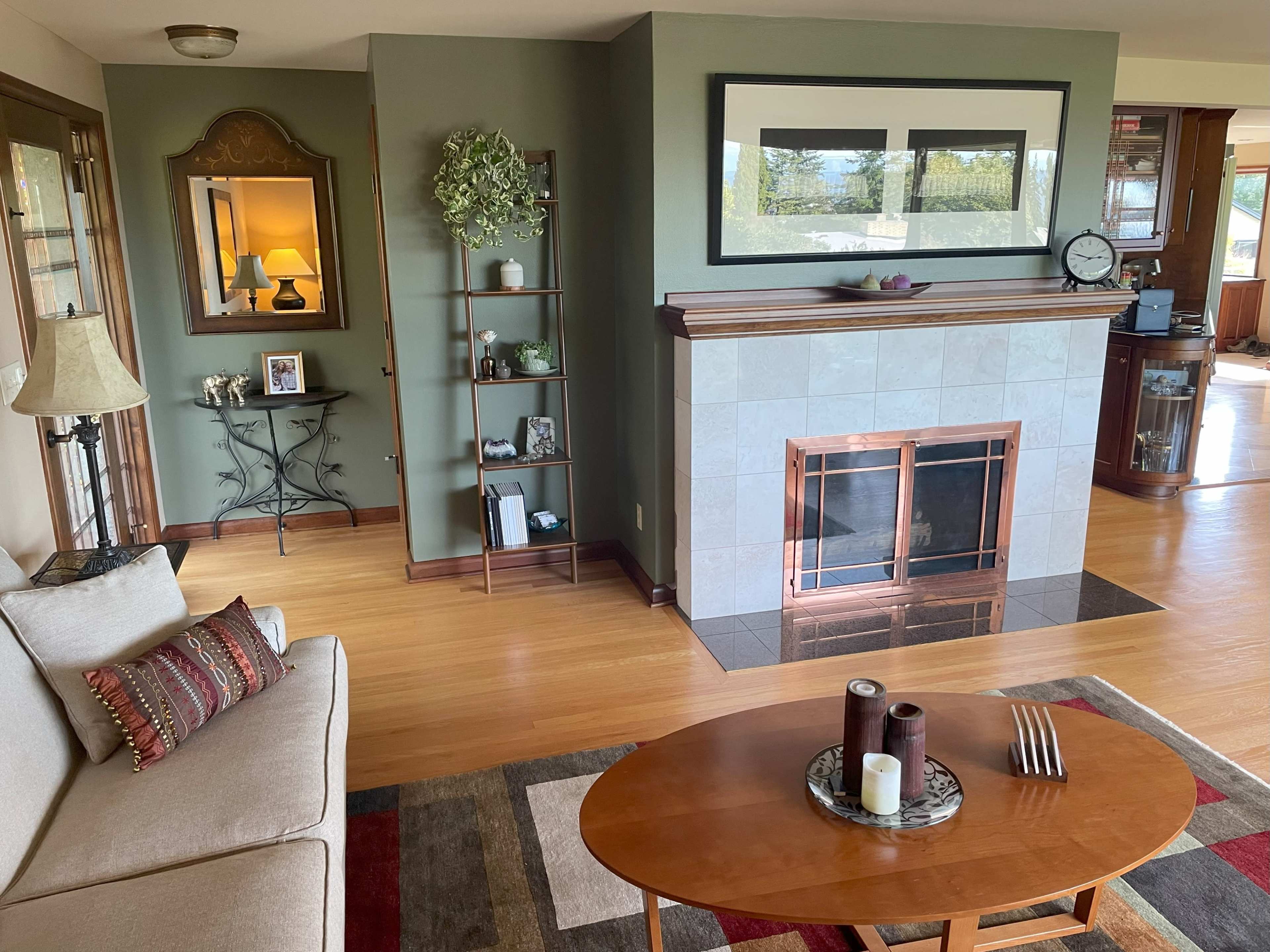 A living room with a beige sofa, a wooden coffee table, a fireplace with a copper trim, and a shelf displaying decorative items.