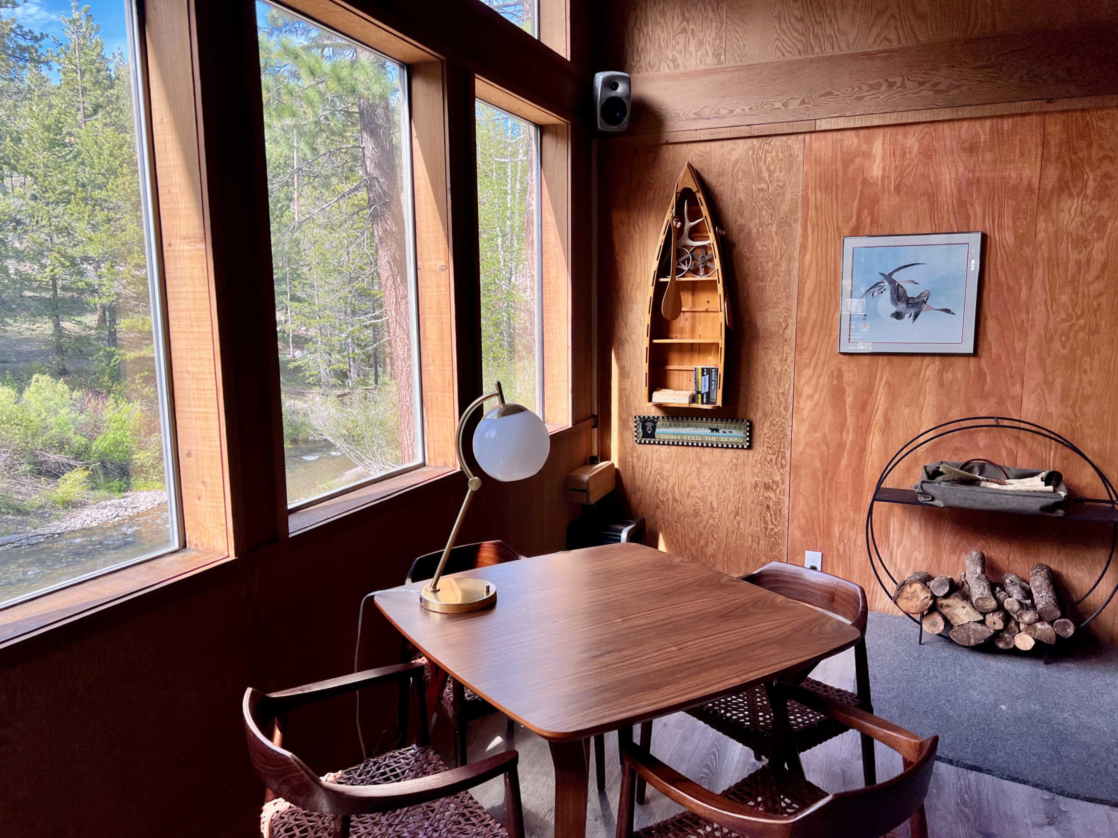 A small wooden table with chairs is positioned near large windows overlooking a wooded area, complemented by a decorative shelf and a lamp on the table.
