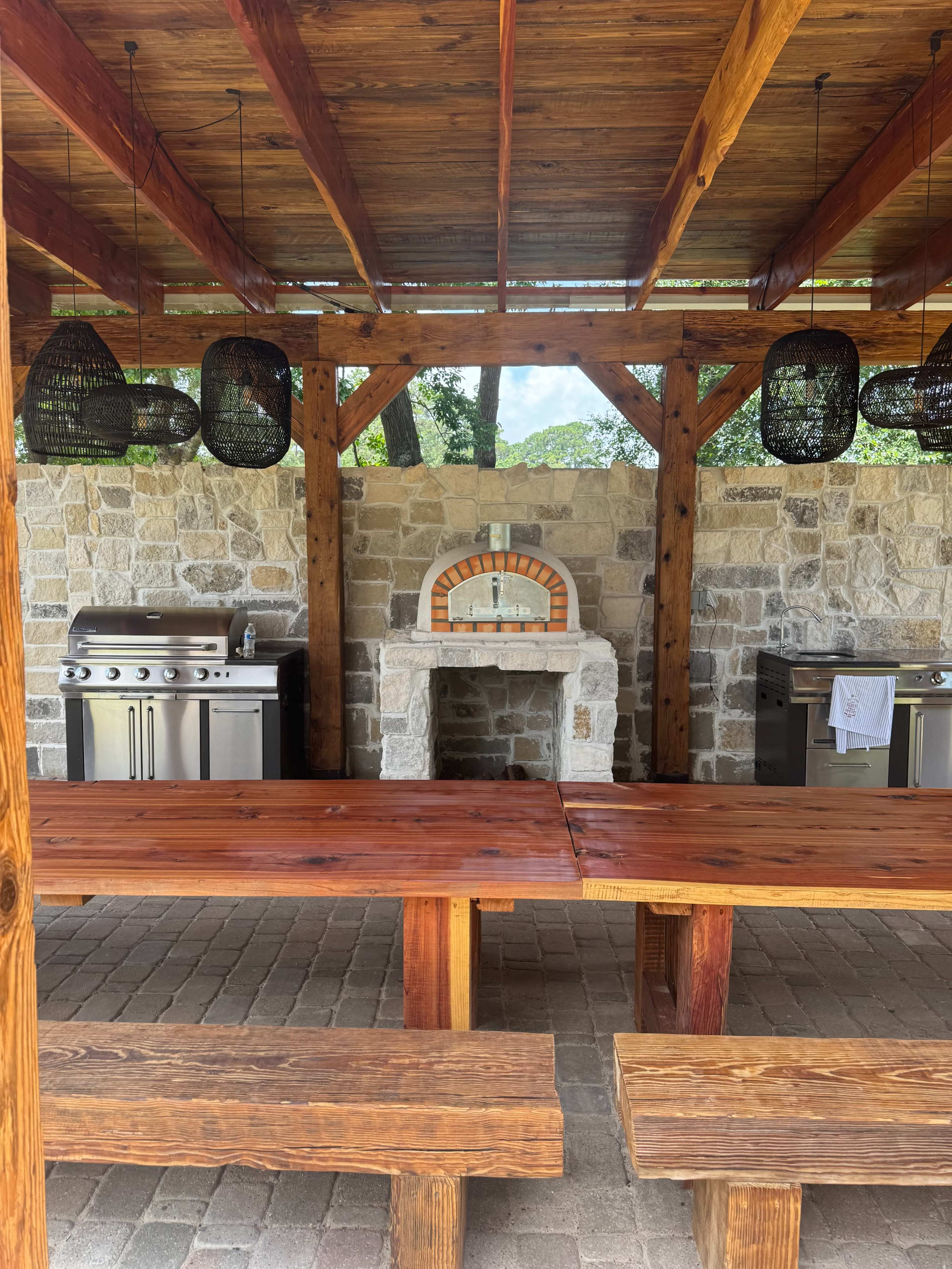 The image depicts an outdoor kitchen area with stone walls, wooden beams, a pizza oven, and two grills, accompanied by a large wooden dining table and benches.