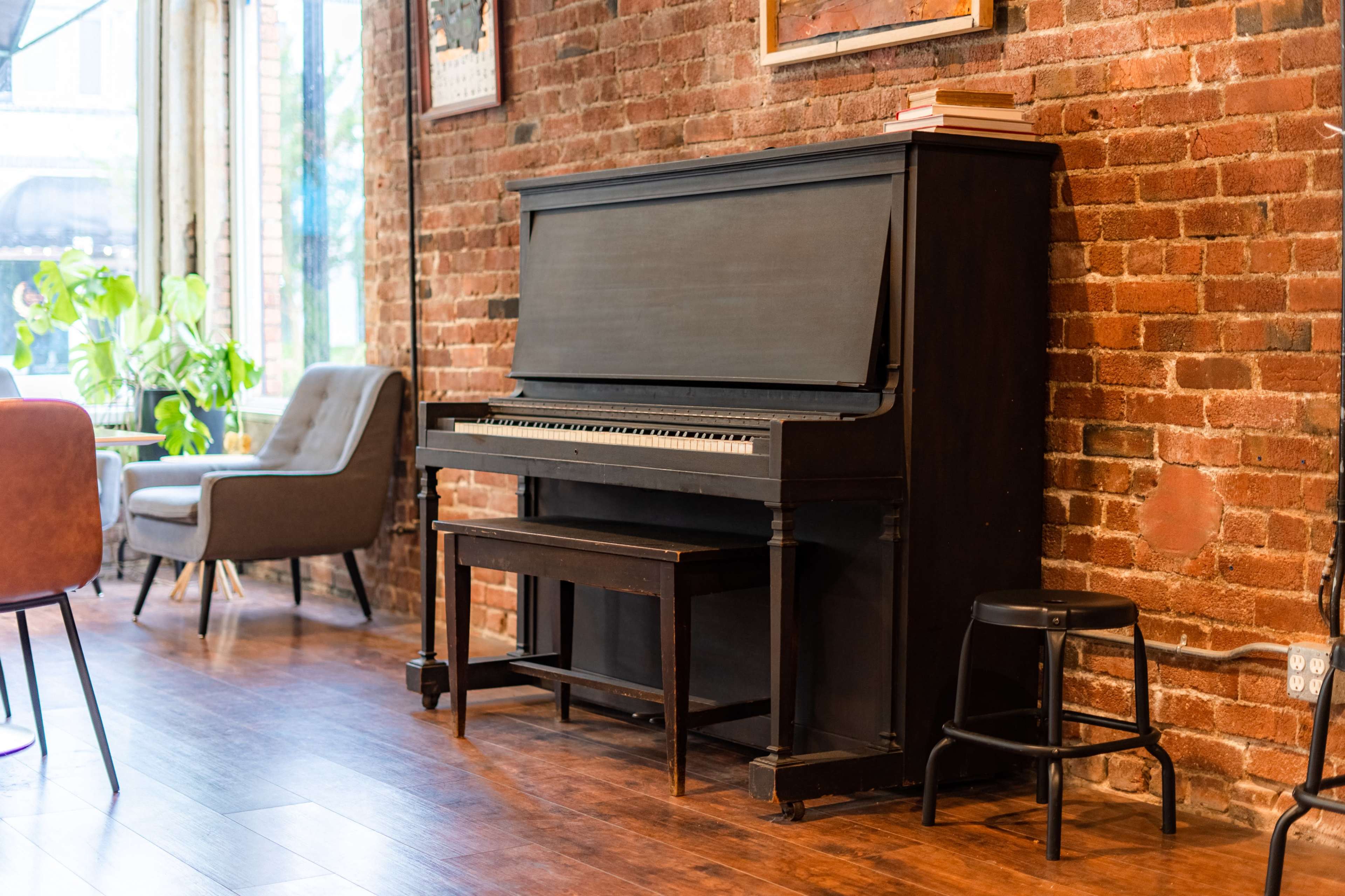 A black upright piano is positioned against a brick wall in a cozy interior space, accompanied by a stool and a few books placed on top.