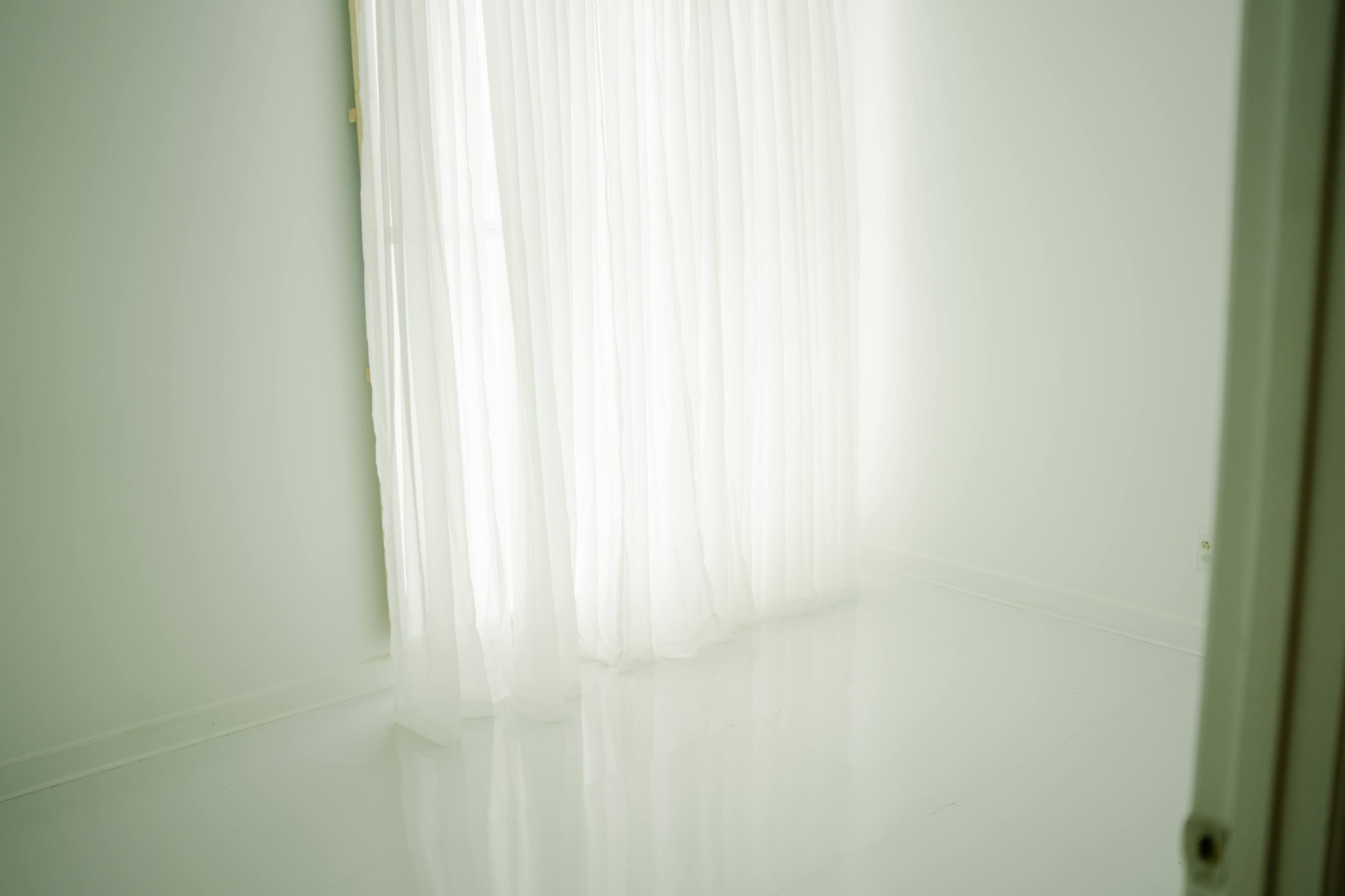 A bright, minimalistic room features sheer white curtains that diffuse natural light against a glossy white floor.