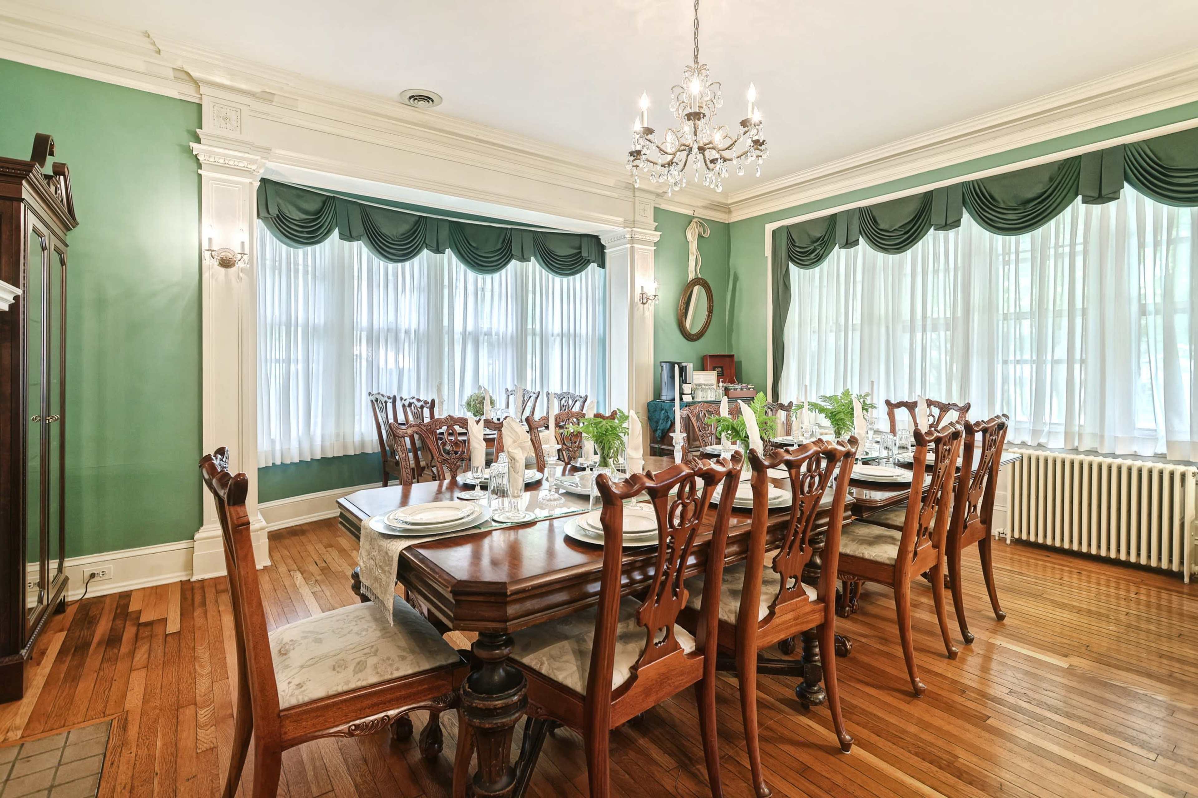 A long dining table with elegant wooden chairs is set for a meal in a green-walled dining room featuring large windows and decorative curtains.