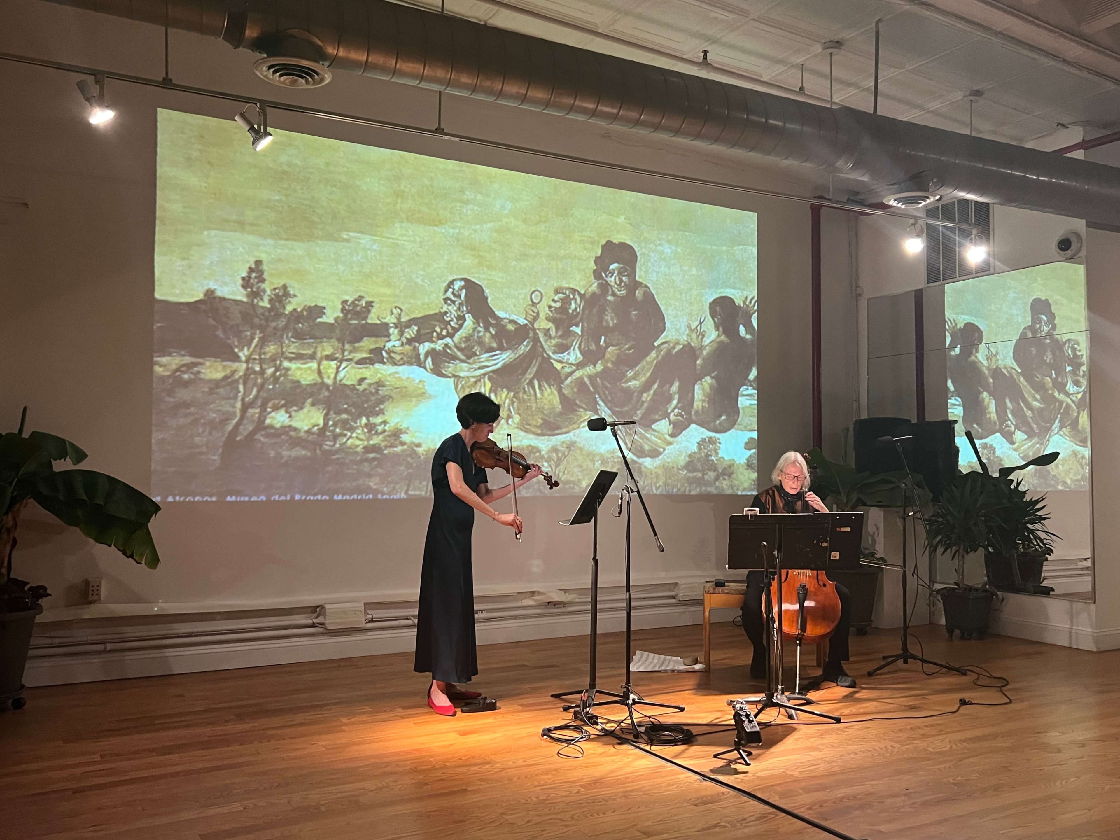 A woman plays the violin while another musician sits beside her with a cello, in front of a large screen displaying an artwork.