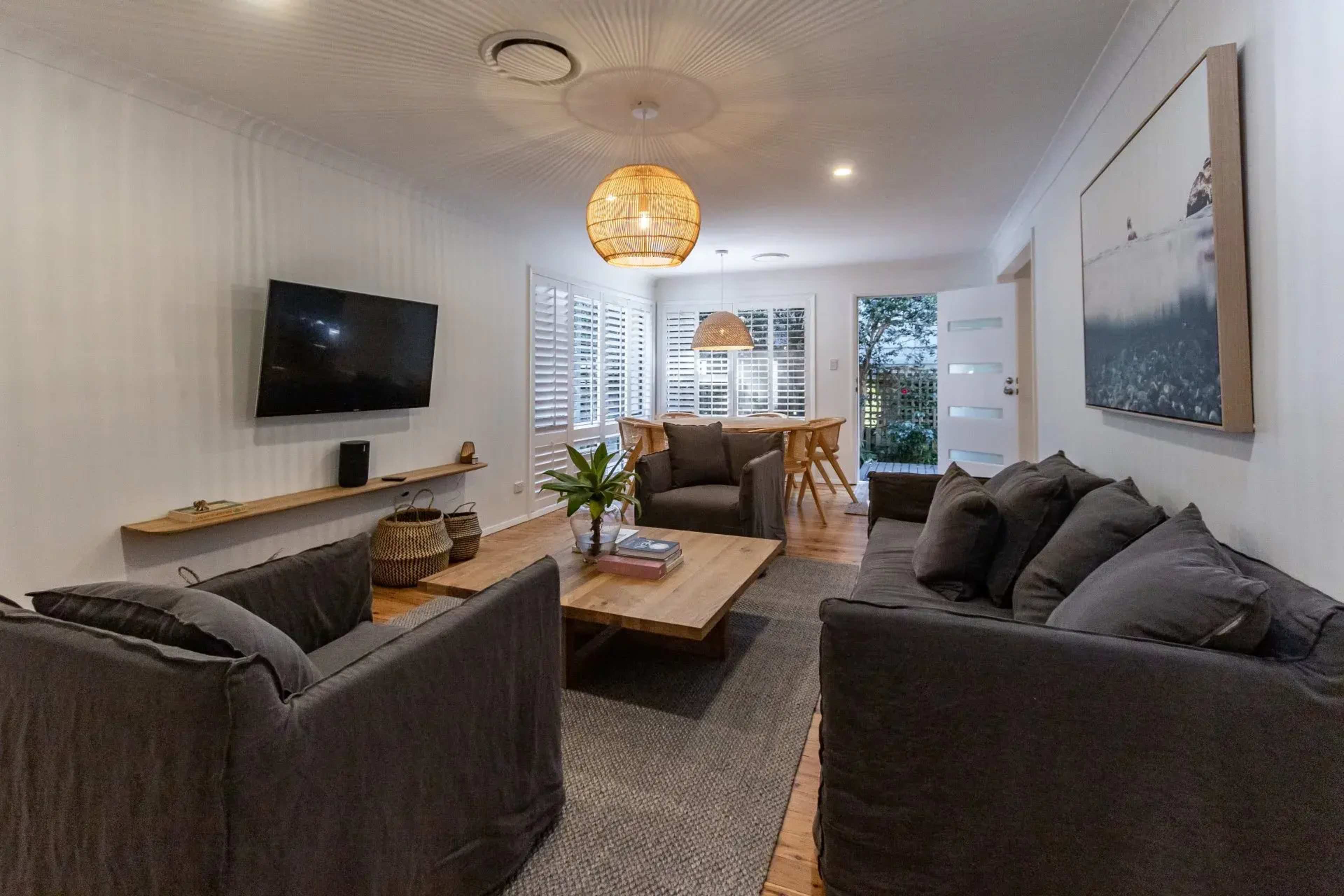 The room features two dark sofas facing a wooden coffee table, a mounted TV, and a dining area with a round table, illuminated by a woven pendant light.
