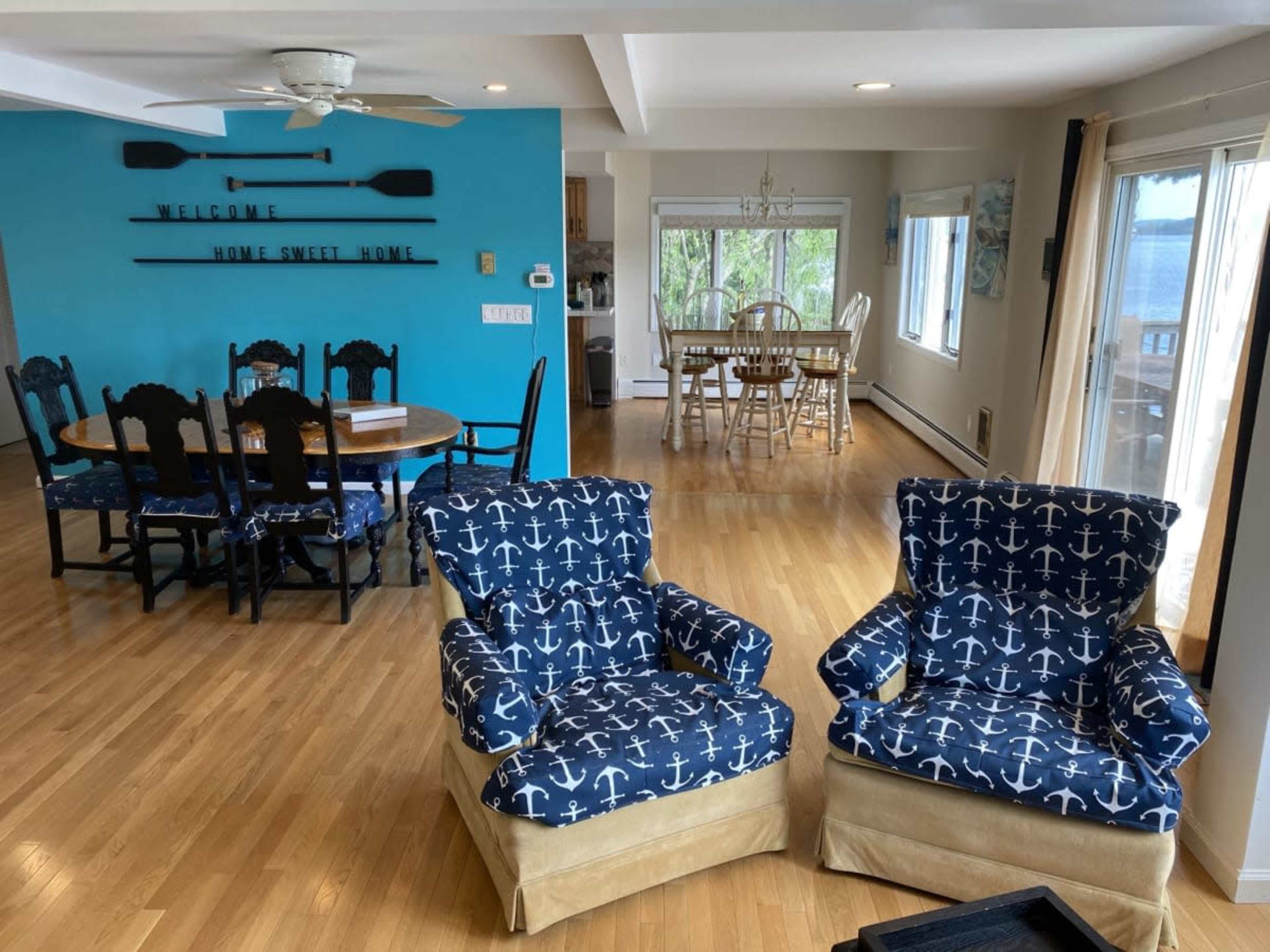 A bright interior space featuring two blue upholstered chairs with anchor patterns, a dining table with wooden chairs, and large windows overlooking a body of water.