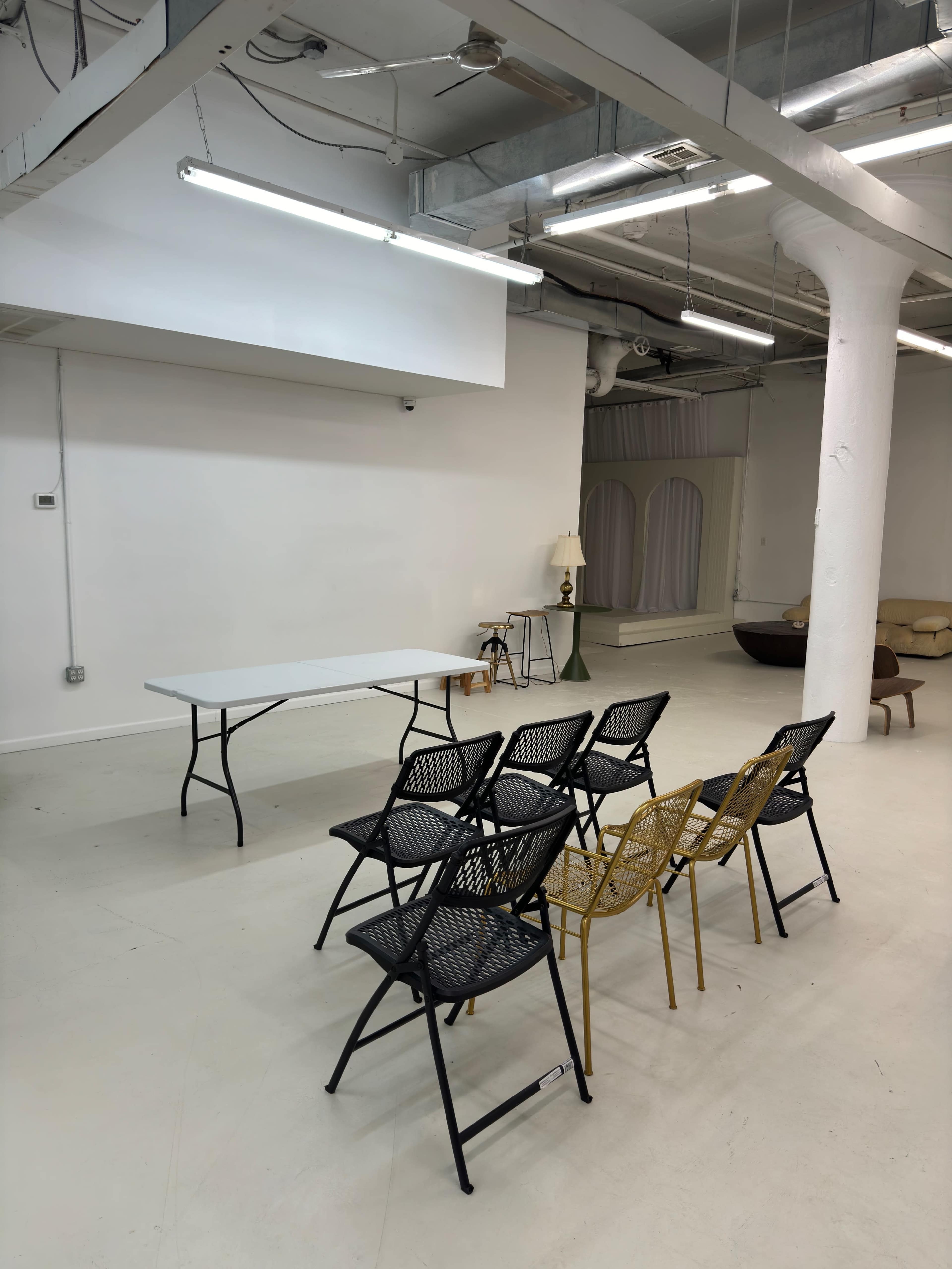 A minimalist room with a row of black and gold chairs facing a white table, illuminated by overhead lights.