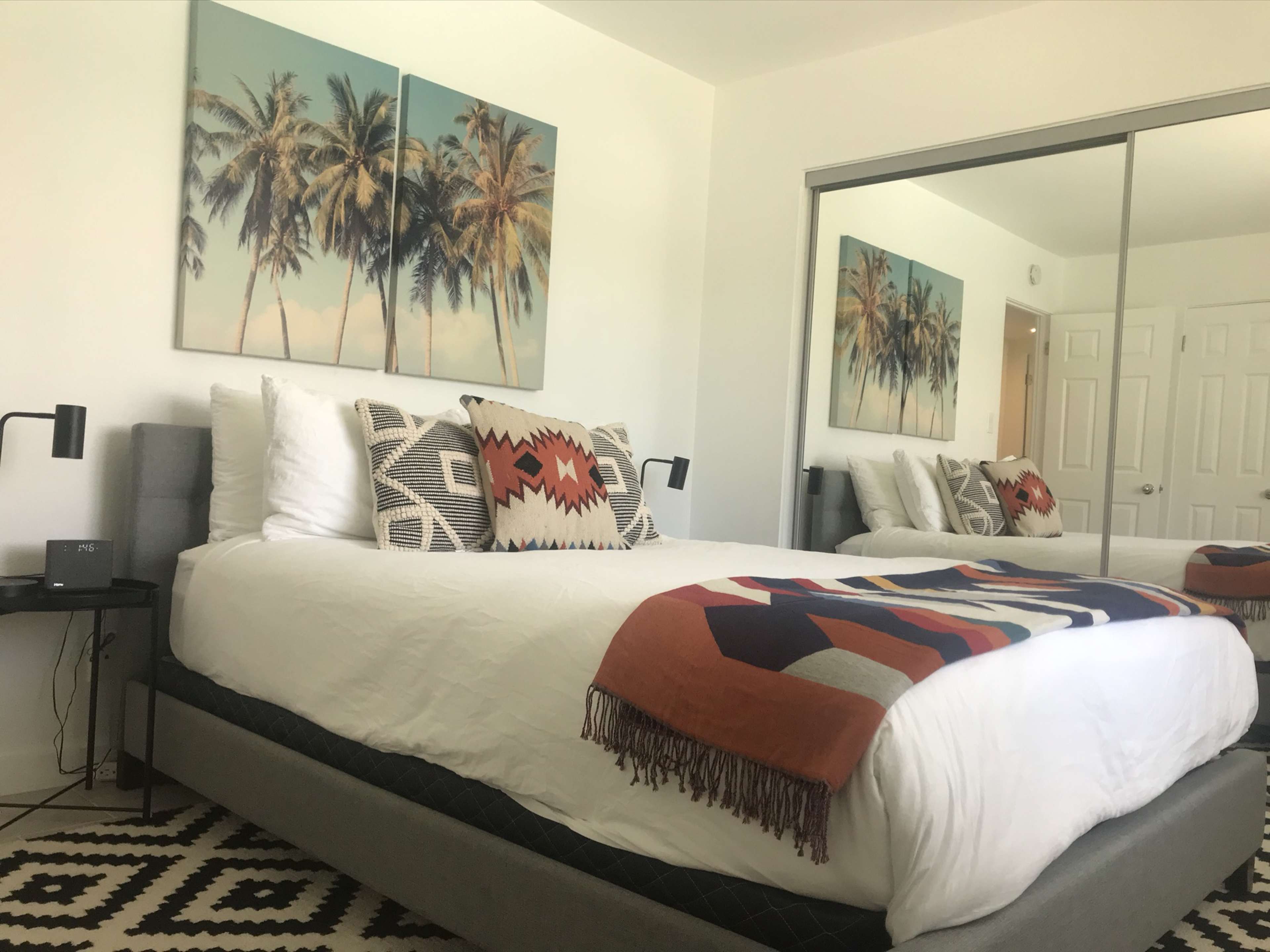 The image shows a neatly arranged bedroom featuring a large bed with decorative pillows, two palm tree paintings on the wall, and a mirrored closet reflecting the room's design.