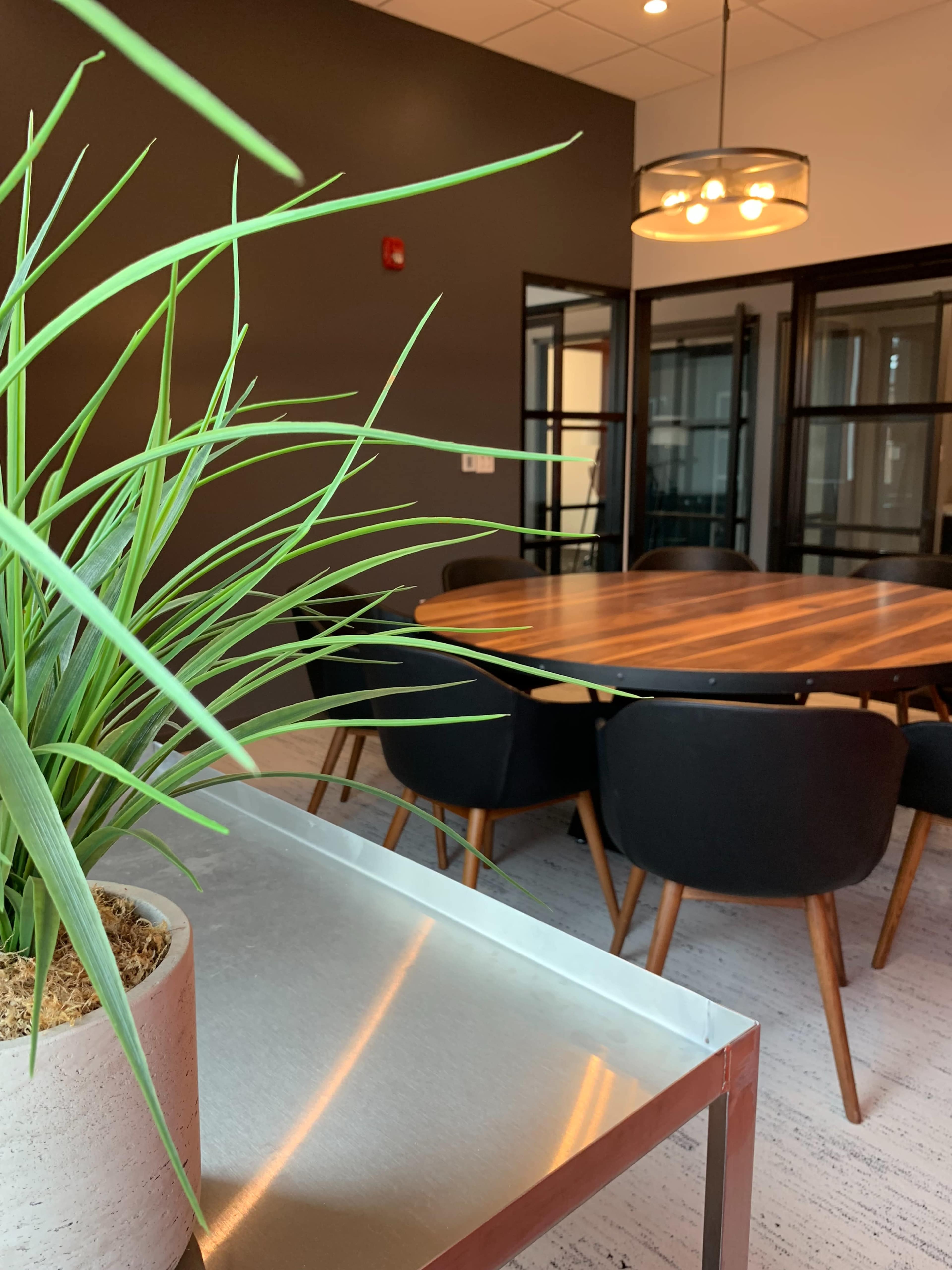 A modern conference room features a large wooden table surrounded by black chairs, with a potted plant visible in the foreground.