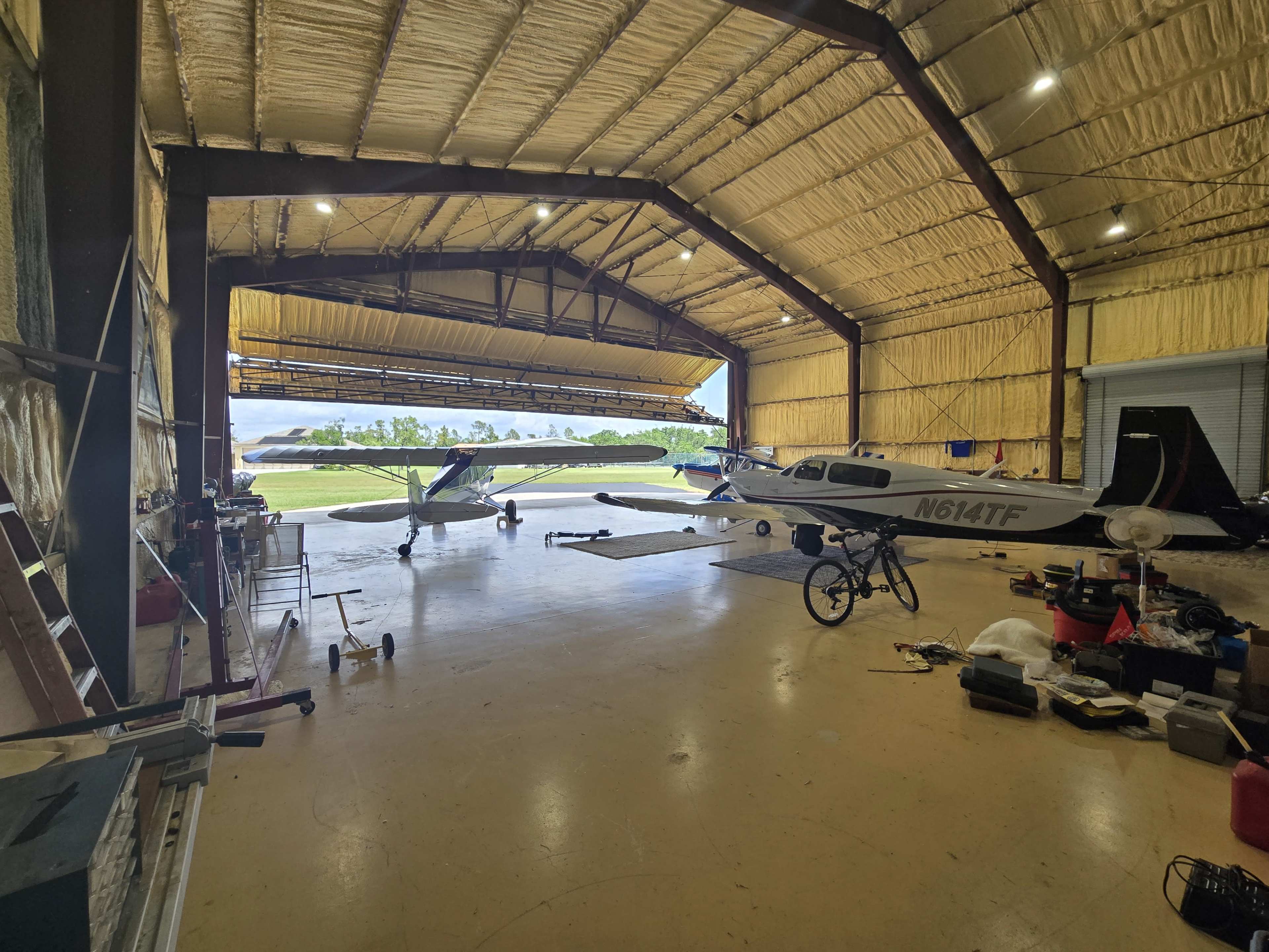 The image shows a spacious hangar with two parked planes, one of which is labeled "N614TF," and various tools and equipment scattered on the floor.