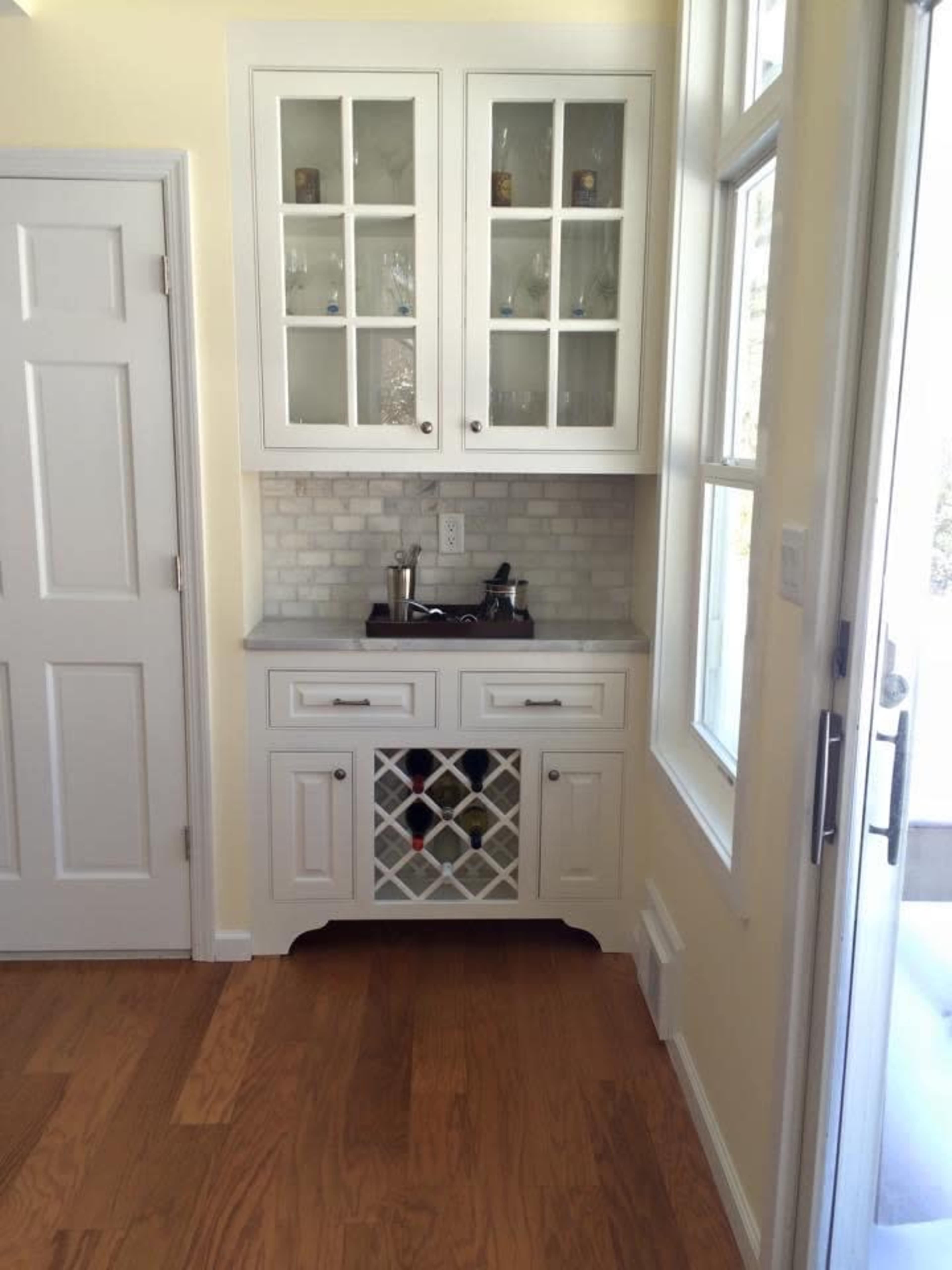 A corner kitchen area with white cabinets featuring glass doors, a wine rack below, and light-colored walls and wooden flooring.