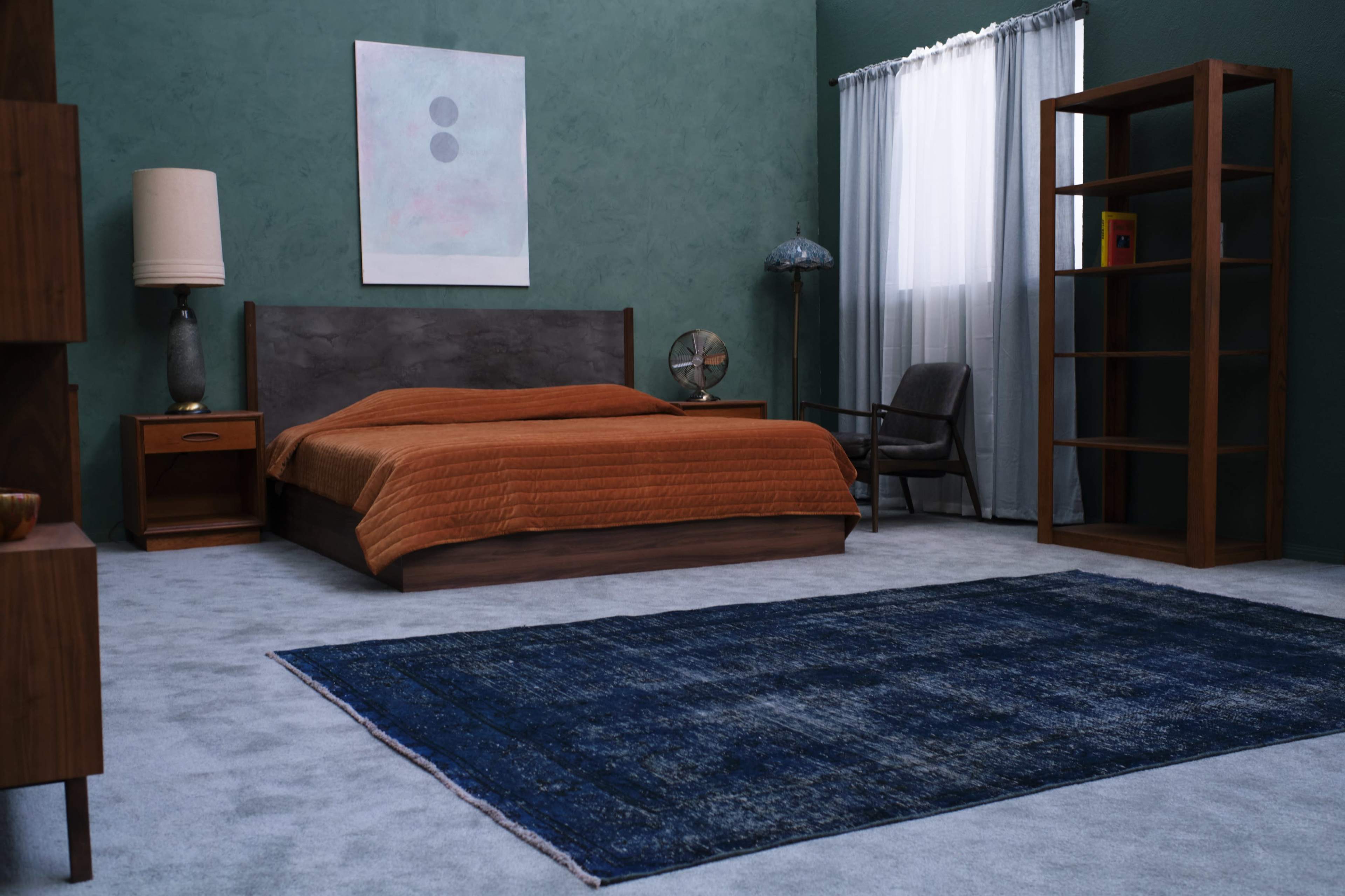 A minimalist bedroom with a wooden bed, an orange bedspread, a gray carpet, and a window with sheer curtains.