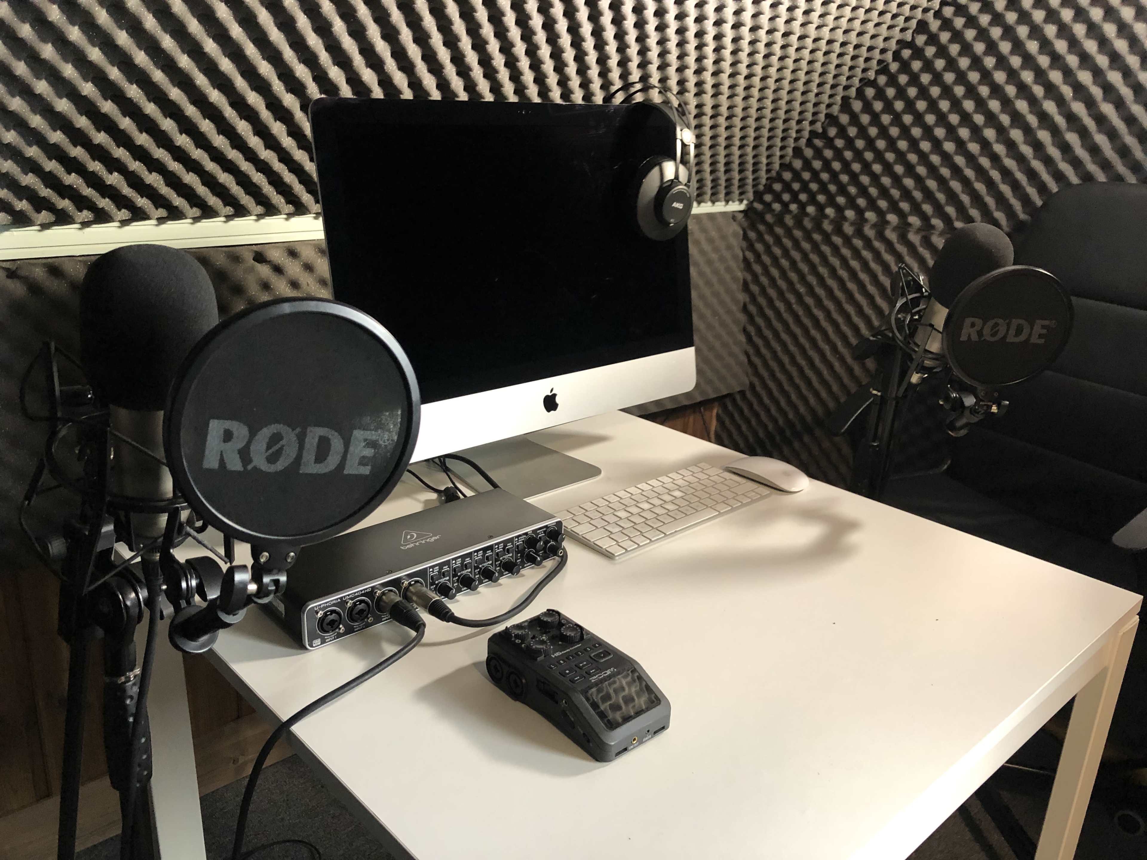 The image shows a recording studio setup featuring two Rode microphones, an audio interface, a computer, a keyboard, and a pair of headphones, all placed on a white table in a soundproofed room.