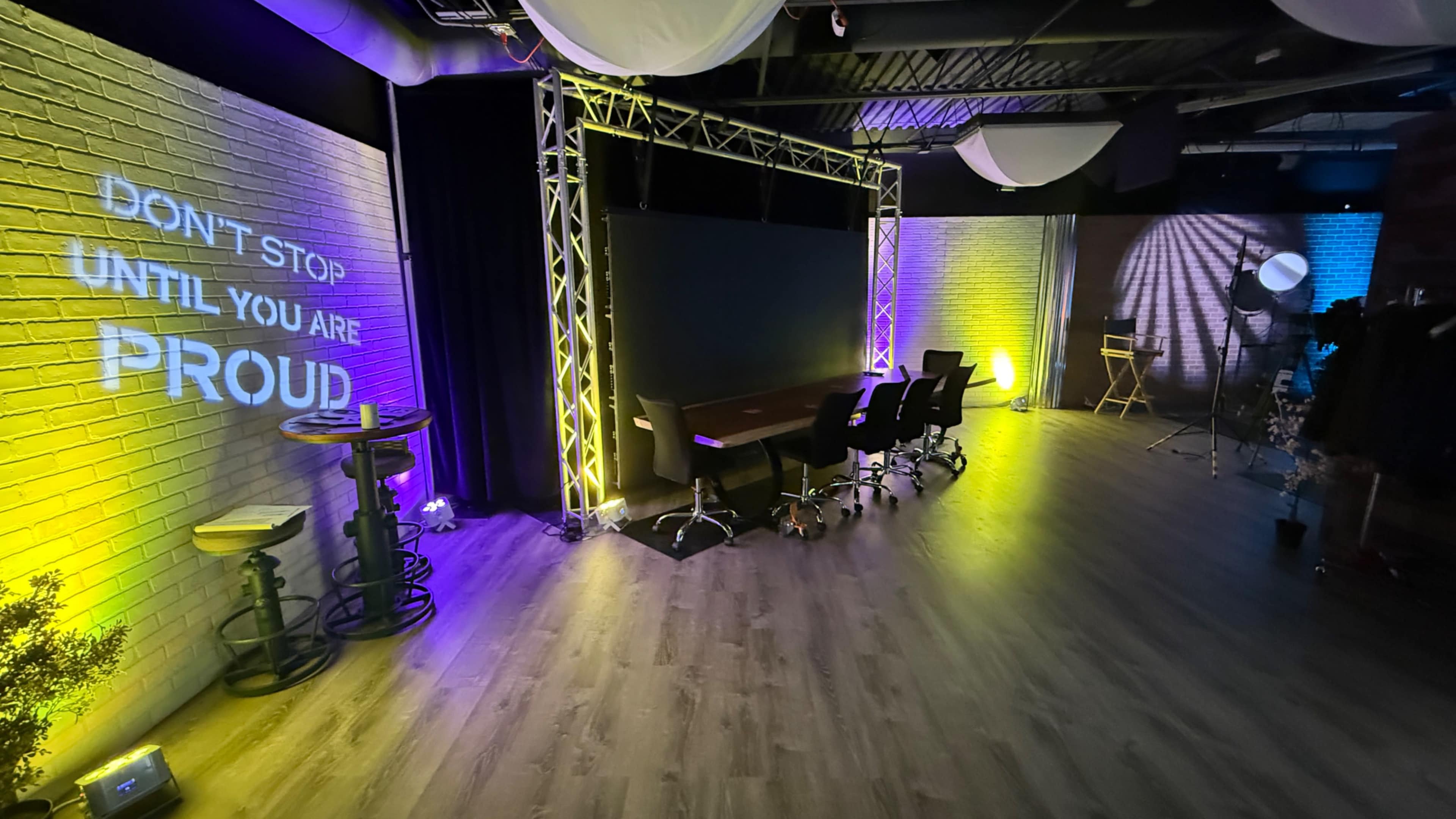 The image shows a modern studio space with a dark backdrop, illuminated walls, and a projection displaying the phrase "DON'T STOP UNTIL YOU ARE PROUD."