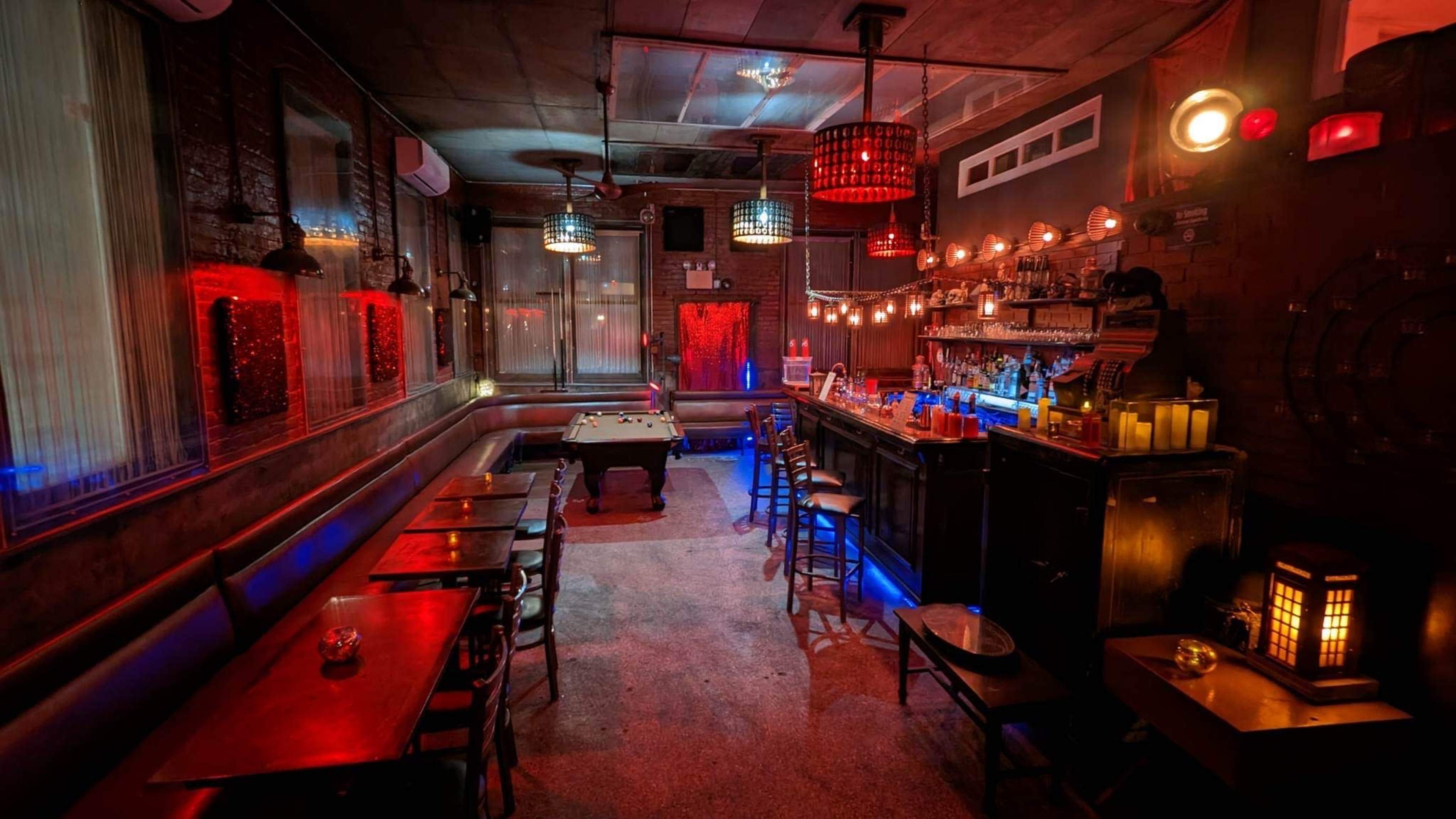 The image shows a dimly lit bar with a pool table, several tables, and a well-stocked bar featuring illuminated bottles.