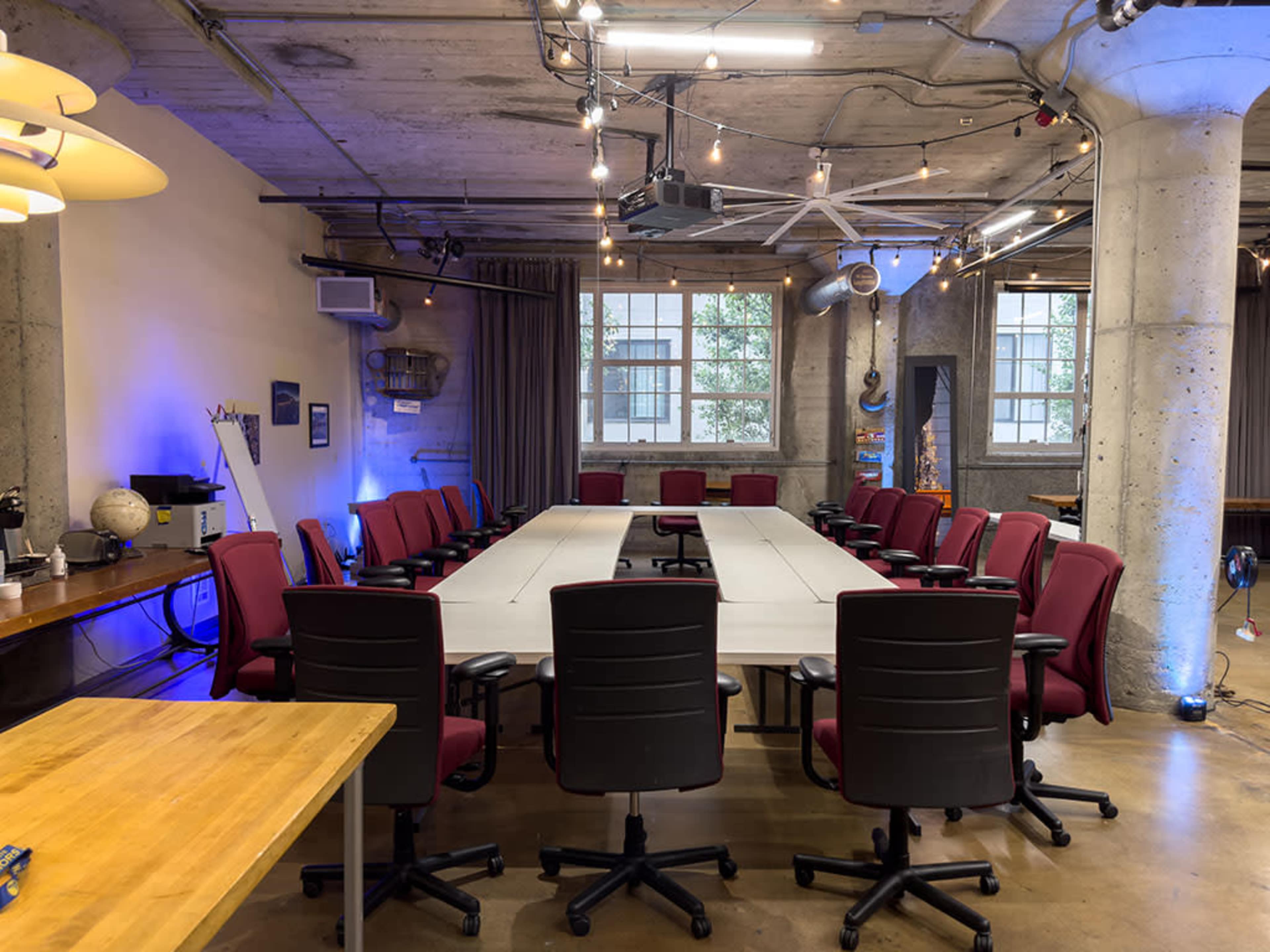 A long, rectangular conference table with red chairs is set up in a modern meeting room featuring exposed concrete walls and industrial lighting.