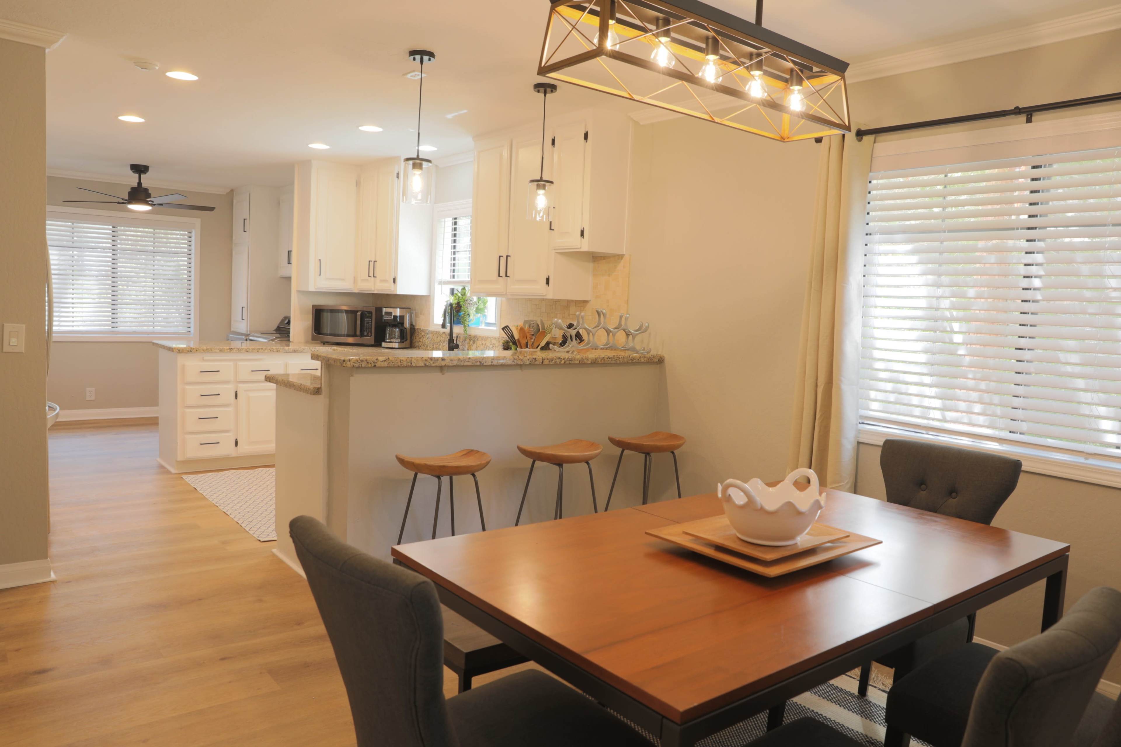 A modern dining area features a wooden table with gray chairs, adjacent to a kitchen with white cabinets and a breakfast bar with three stools.