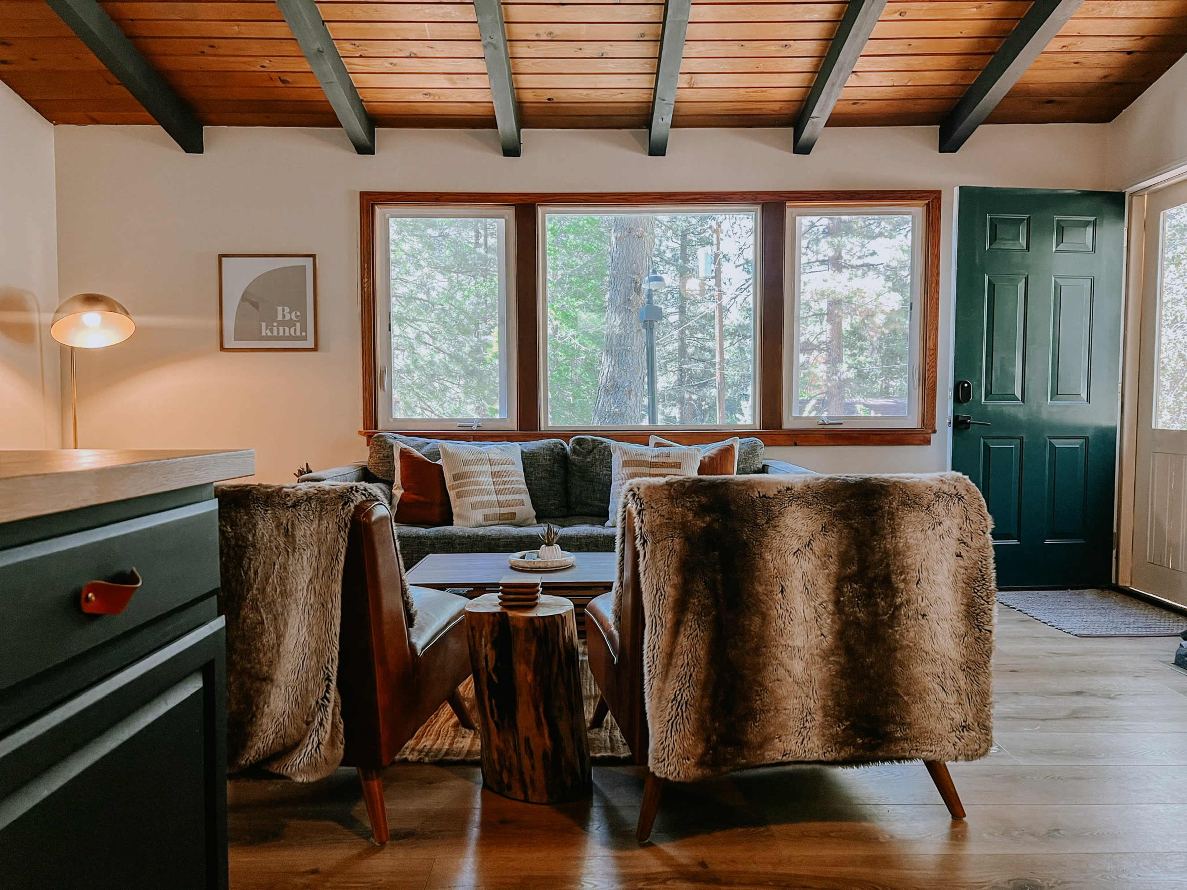 A cozy living room features a gray sofa and two fur-covered chairs, with large windows letting in natural light from the outside.