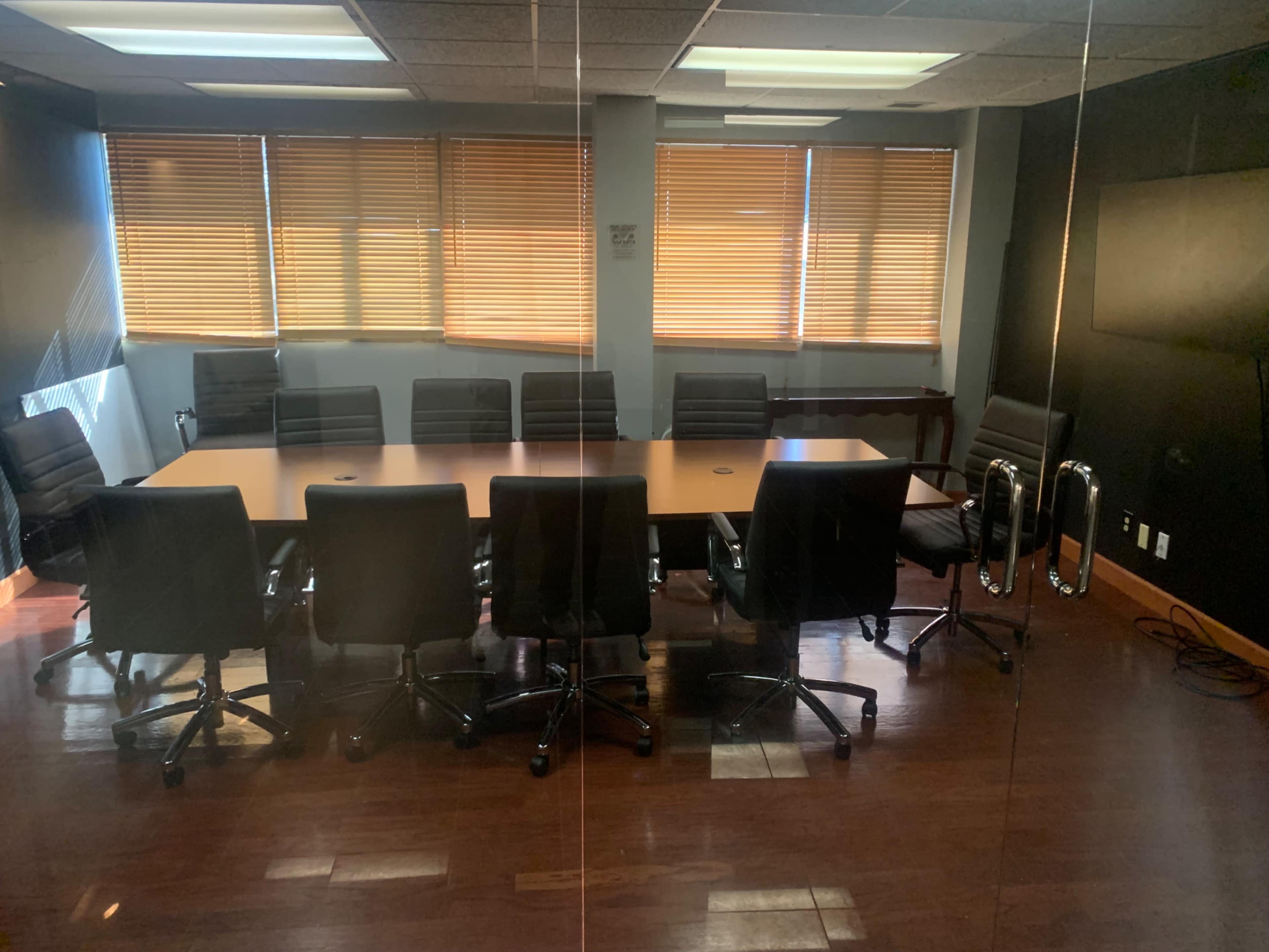 A conference room features a long table surrounded by black office chairs, with windows covered by blinds allowing in warm light.