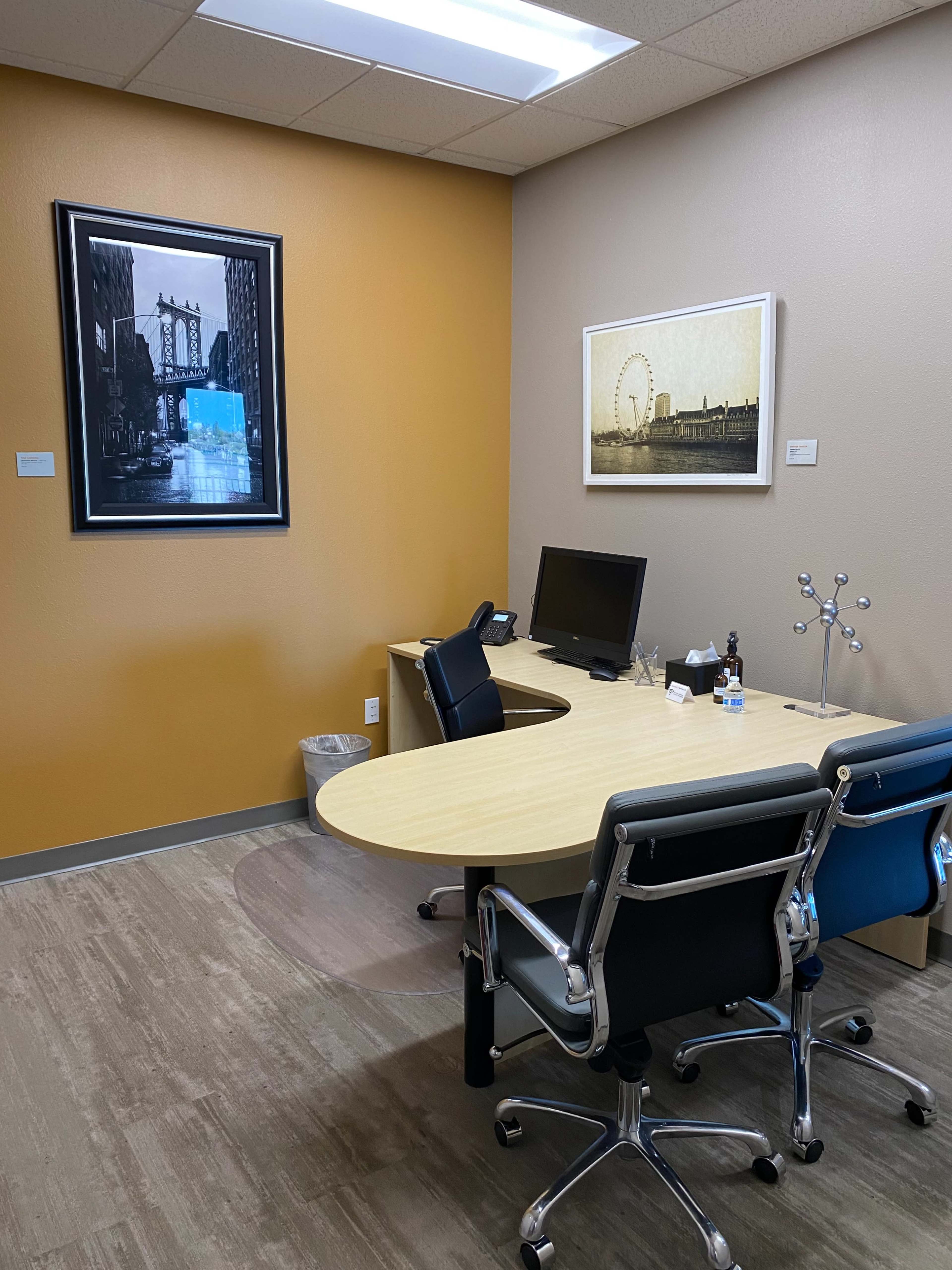 A modern office space with a round wooden desk, two black chairs, a computer monitor, and wall art depicting cityscapes.