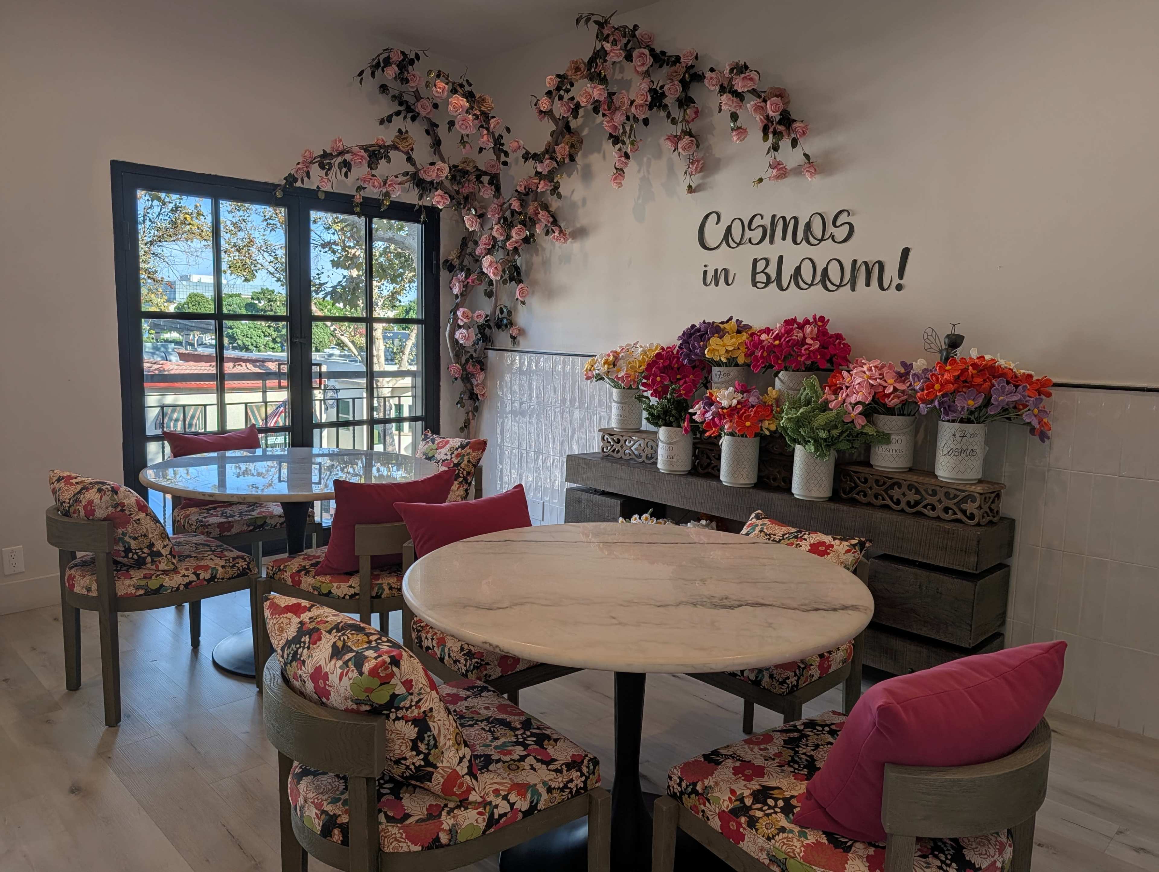 The image shows a stylish cafe interior featuring floral decorations, round marble tables, and colorful upholstered chairs, with a backdrop of a large window and a flower display.