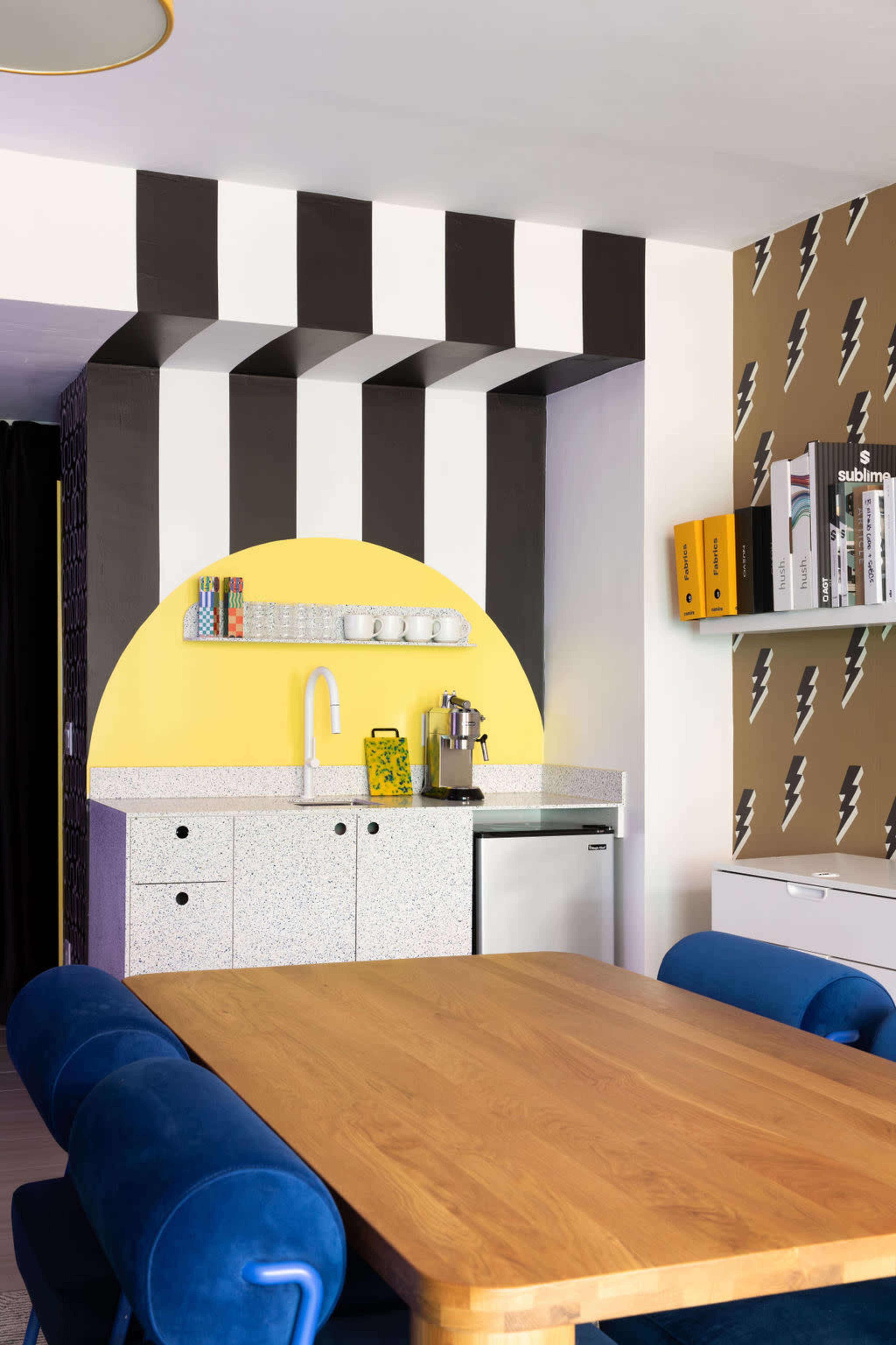 The image shows a modern kitchenette with a yellow semicircular design, black and white striped walls, and a wooden dining table with blue upholstered chairs.