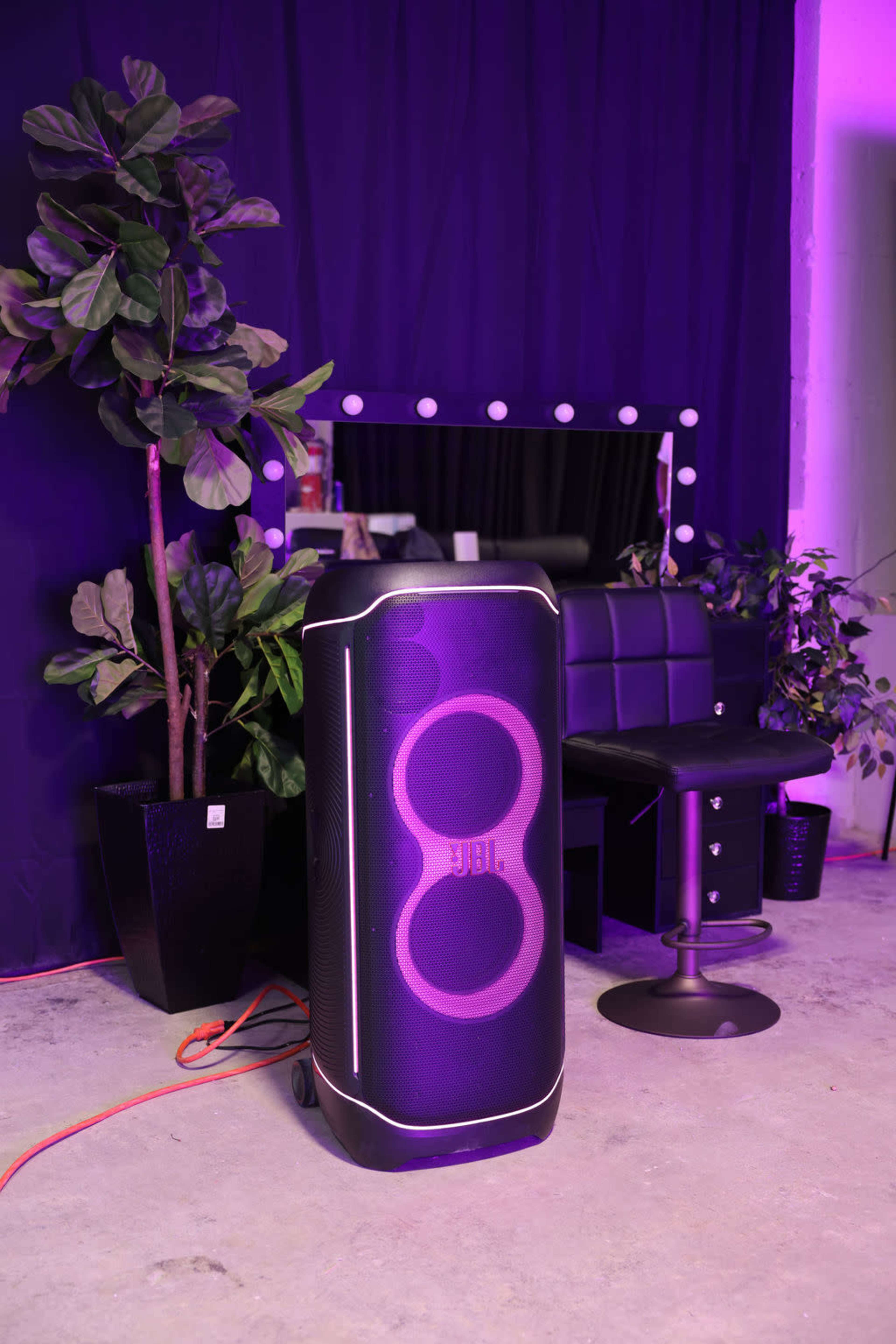 A JBL speaker with a glowing design stands next to a plant and a styling chair in a room with purple lighting.