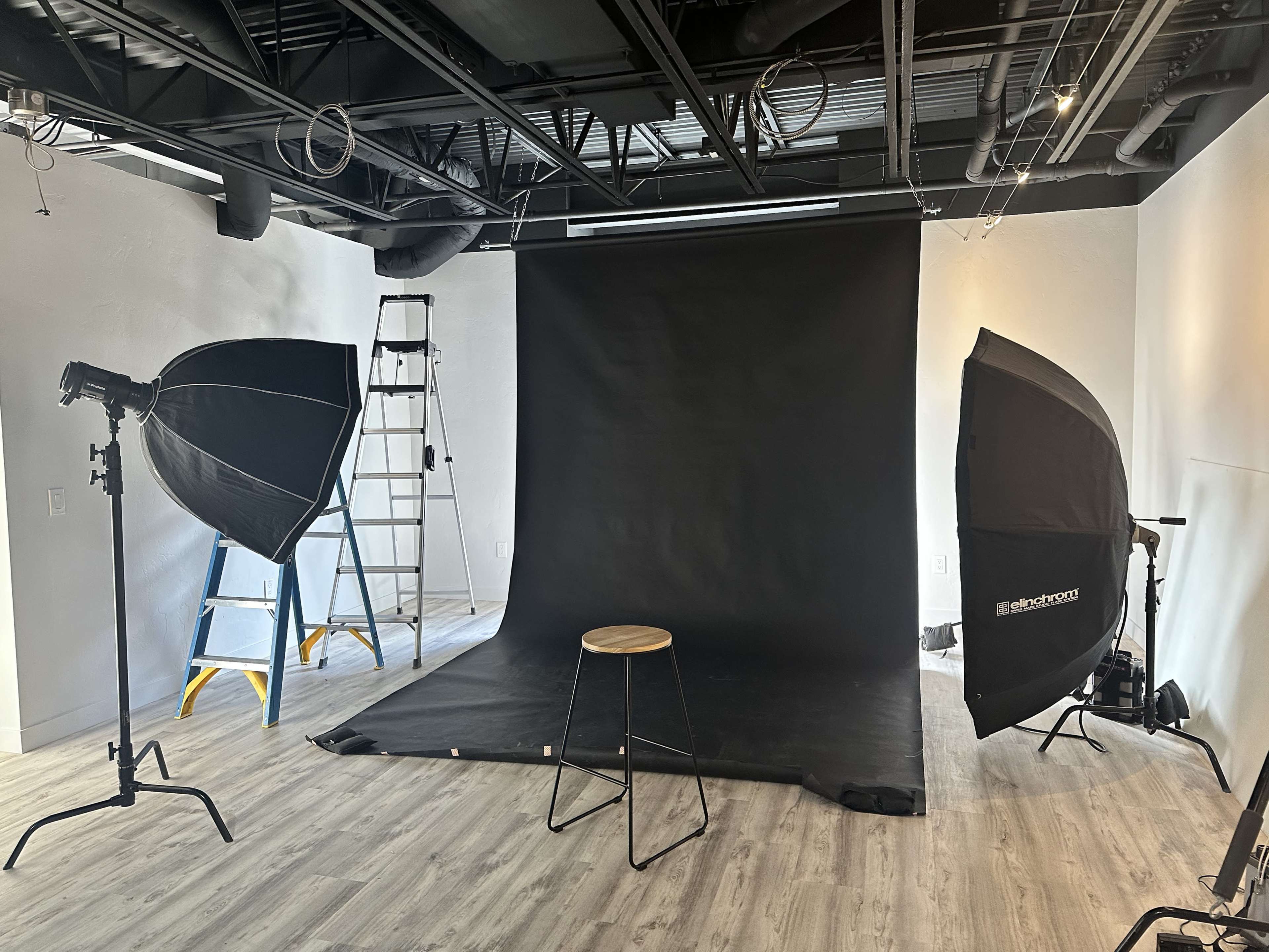 A photo studio setup featuring a black backdrop, a stool in the center, and two lighting softboxes positioned on either side, with a ladder in the background.