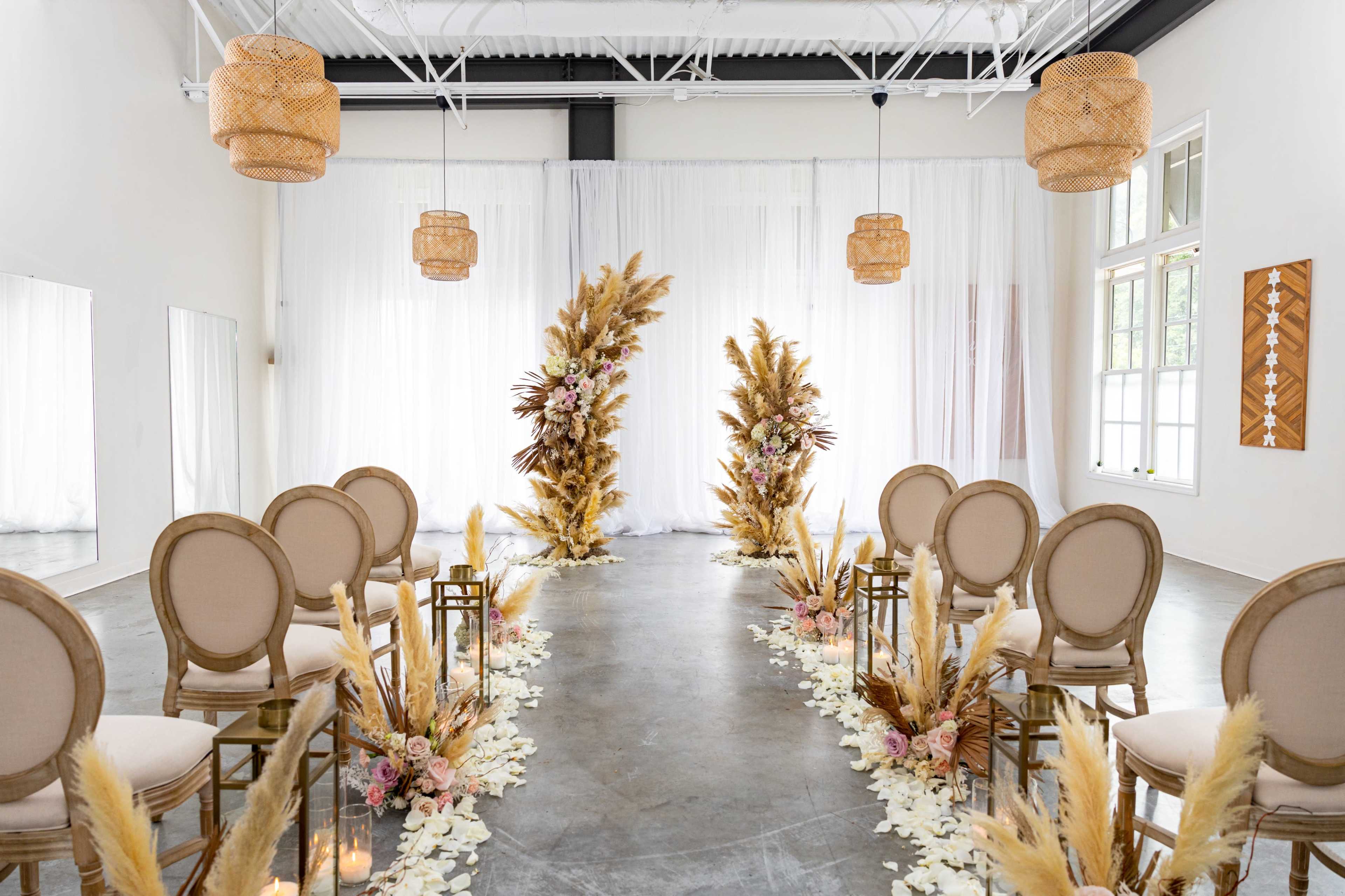 The image shows a wedding ceremony setup featuring rows of chairs flanking a floral arch made of pampas grass and other decorations, all set in a bright, white-walled venue.