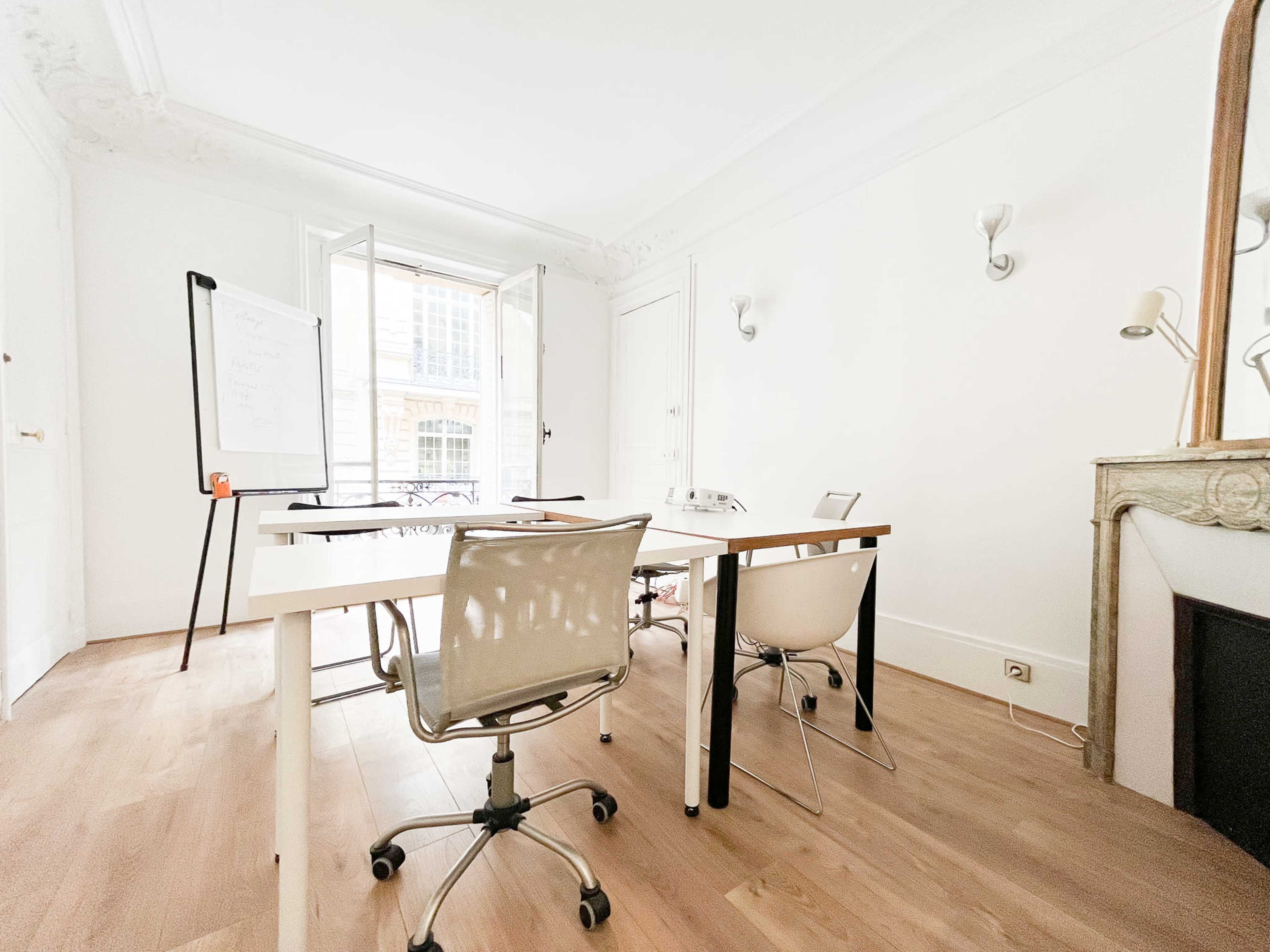 A bright, minimalist office space with a table and several chairs, and large windows opening to an outdoor view.