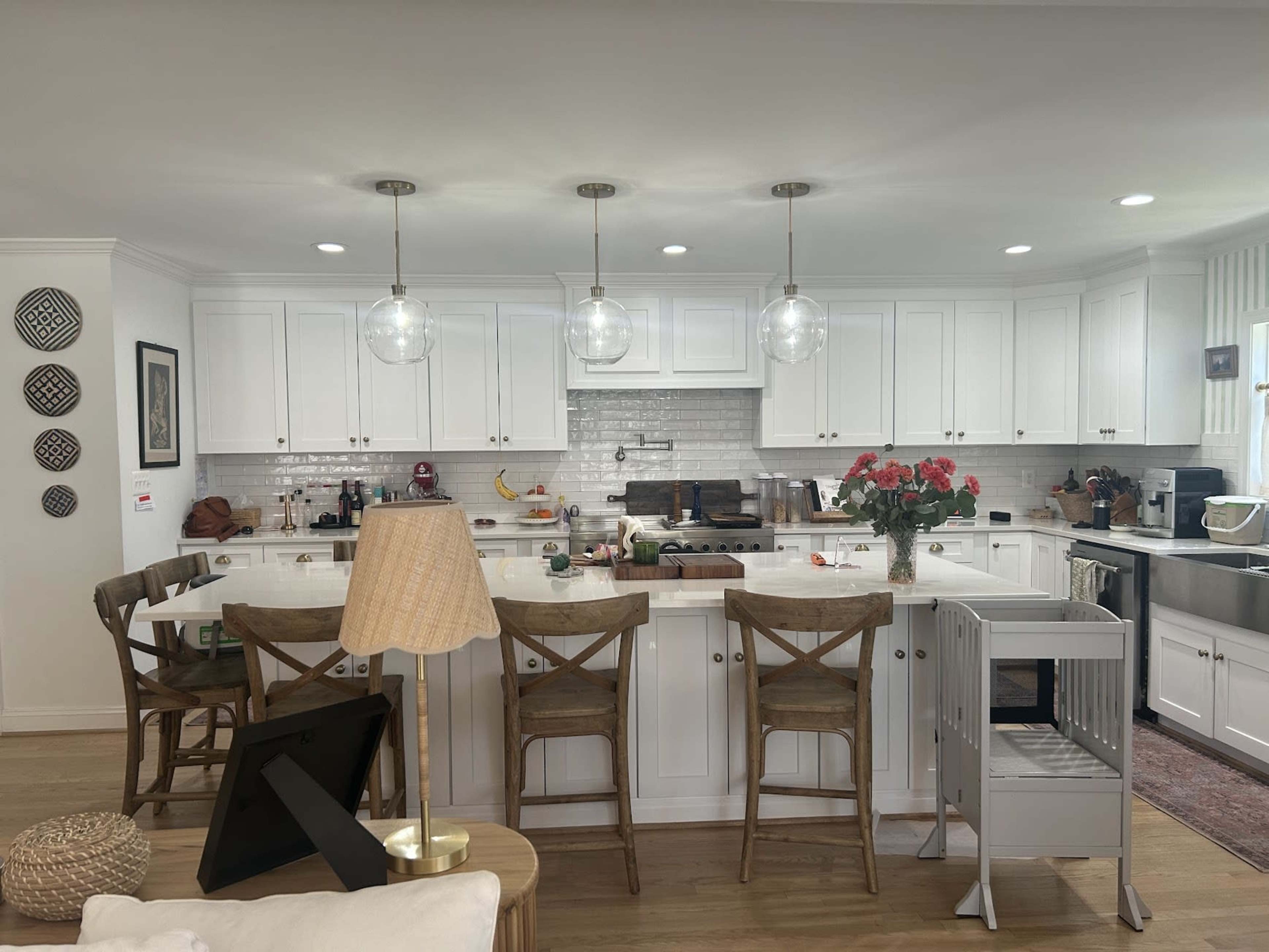 The image shows a modern kitchen with white cabinetry, a central island with bar stools, and pendant lighting, featuring a variety of kitchen appliances and decor items.